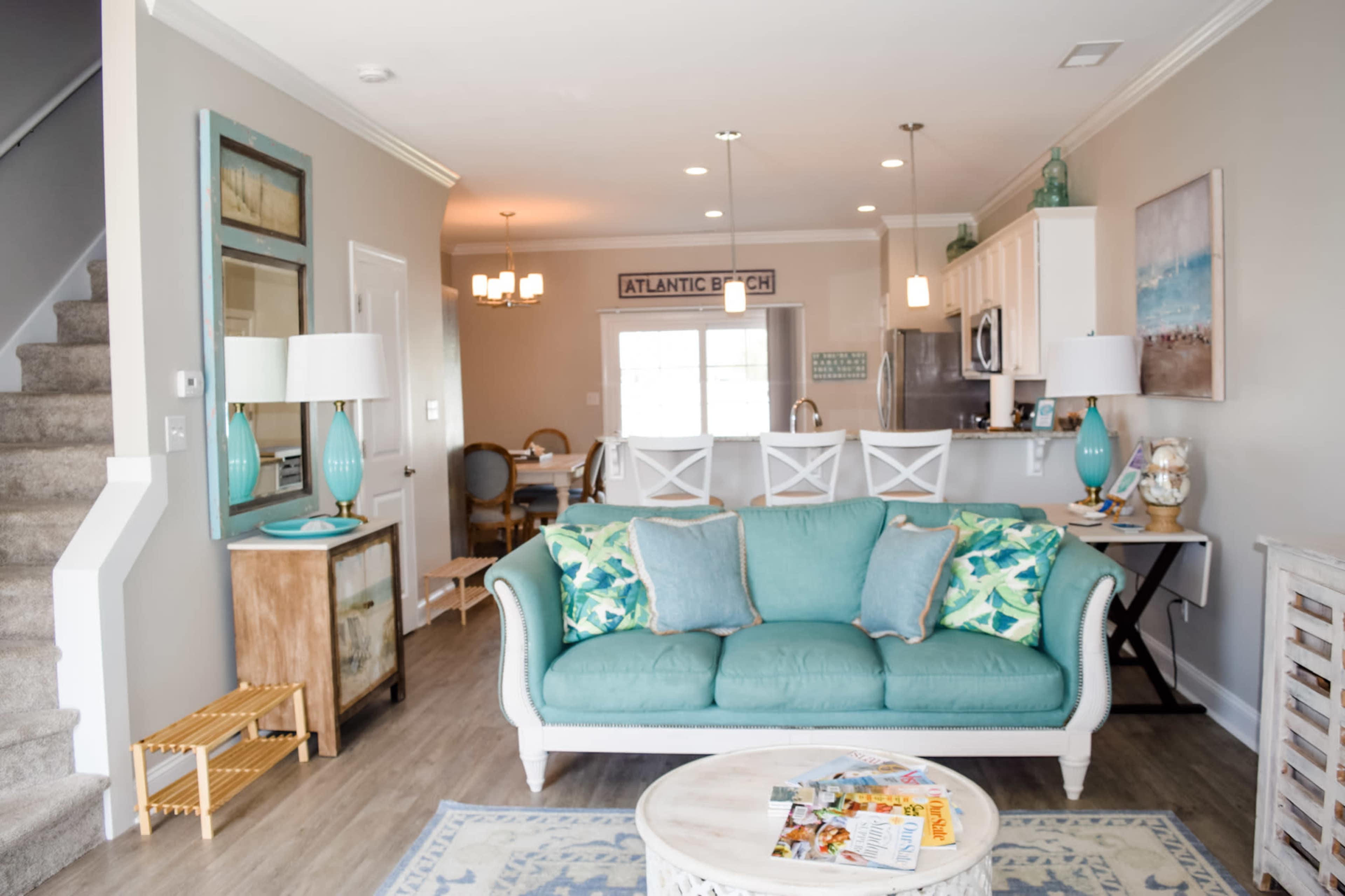 A living area with a turquoise sofa adorned with patterned cushions, a round coffee table, and a dining area visible in the background.