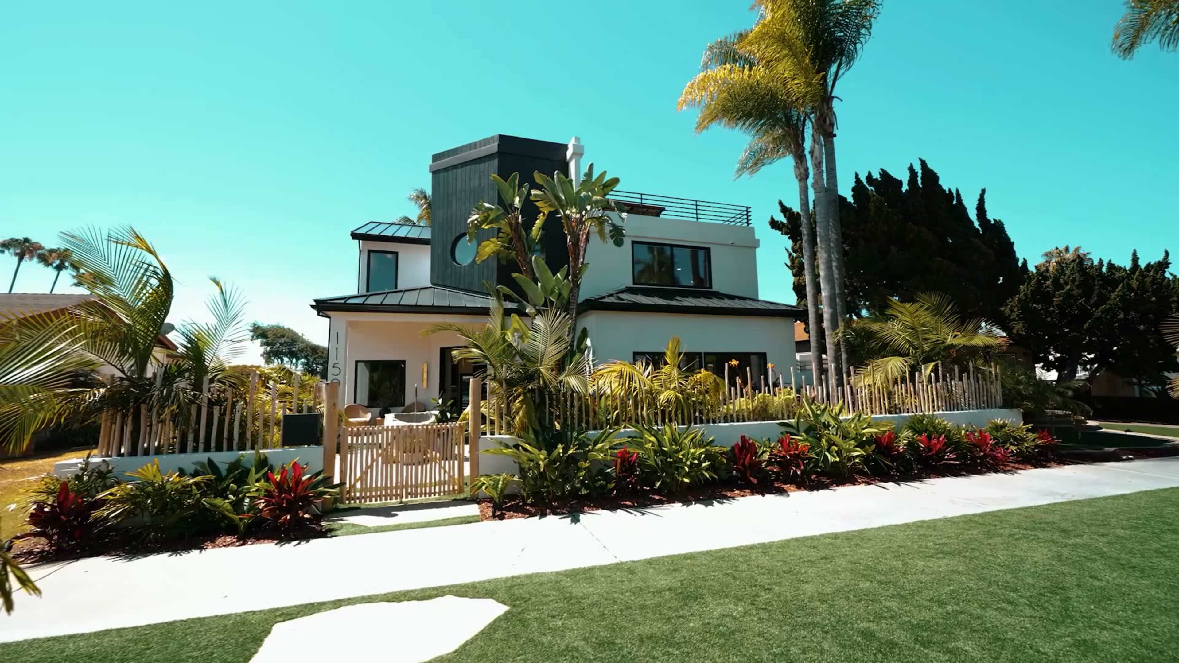 A modern two-story house with a minimalist design is surrounded by lush landscaping and palm trees under a clear blue sky.