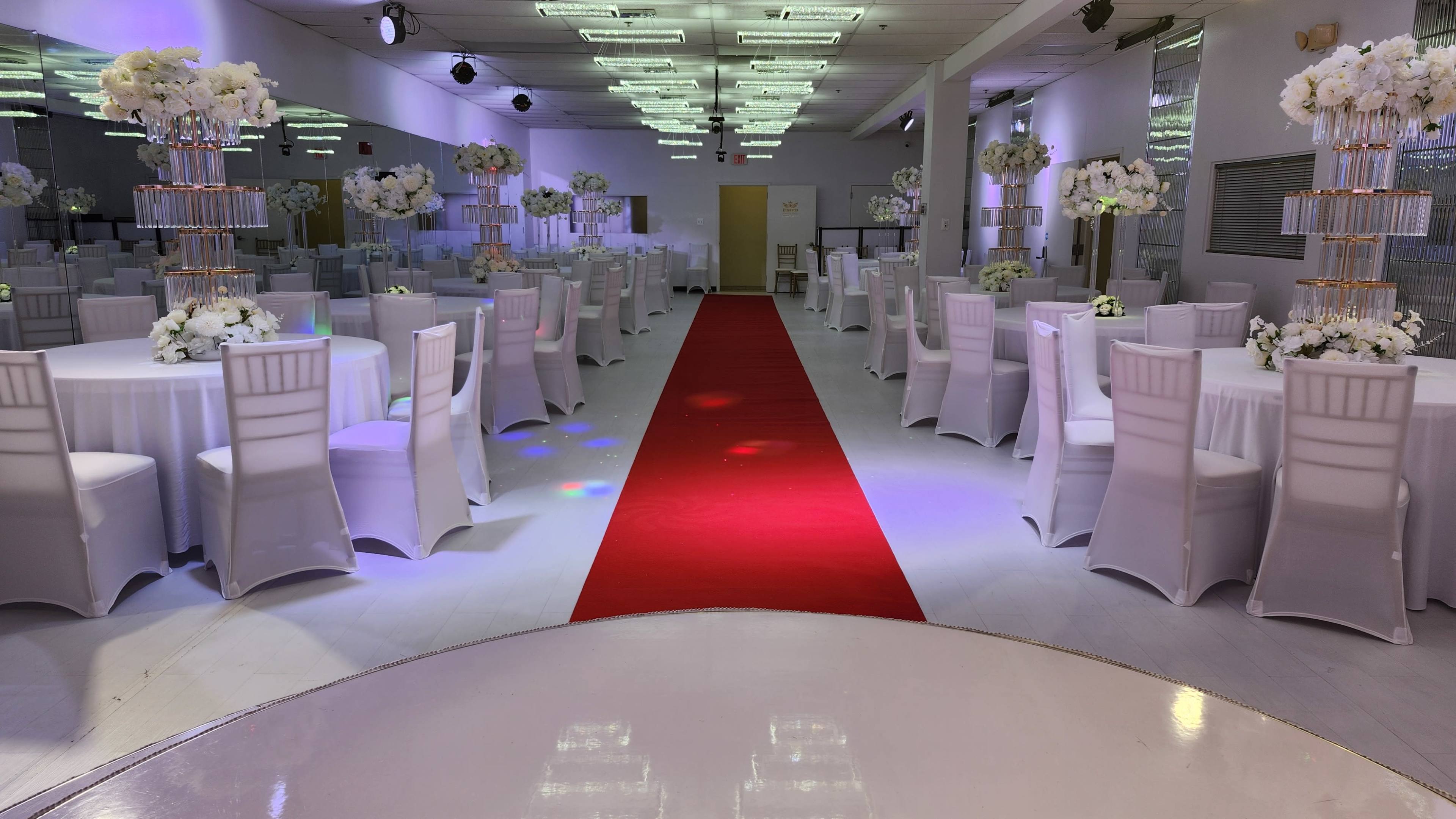 A banquet hall features rows of elegantly set tables with white chairs and floral centerpieces, leading to a red carpet in the center.