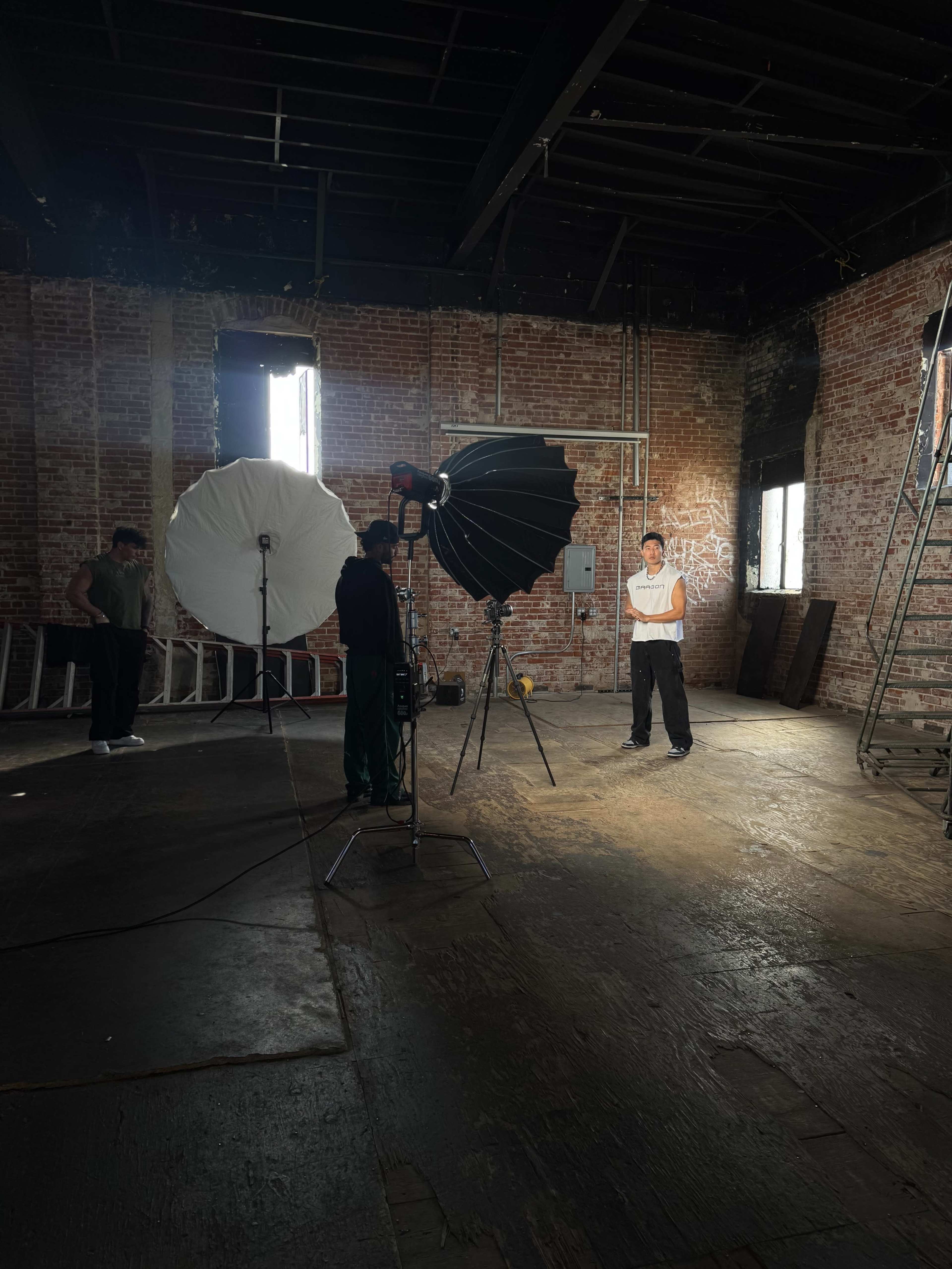 A photographer is setting up lighting equipment in an industrial space while a model stands in front of a backdrop.