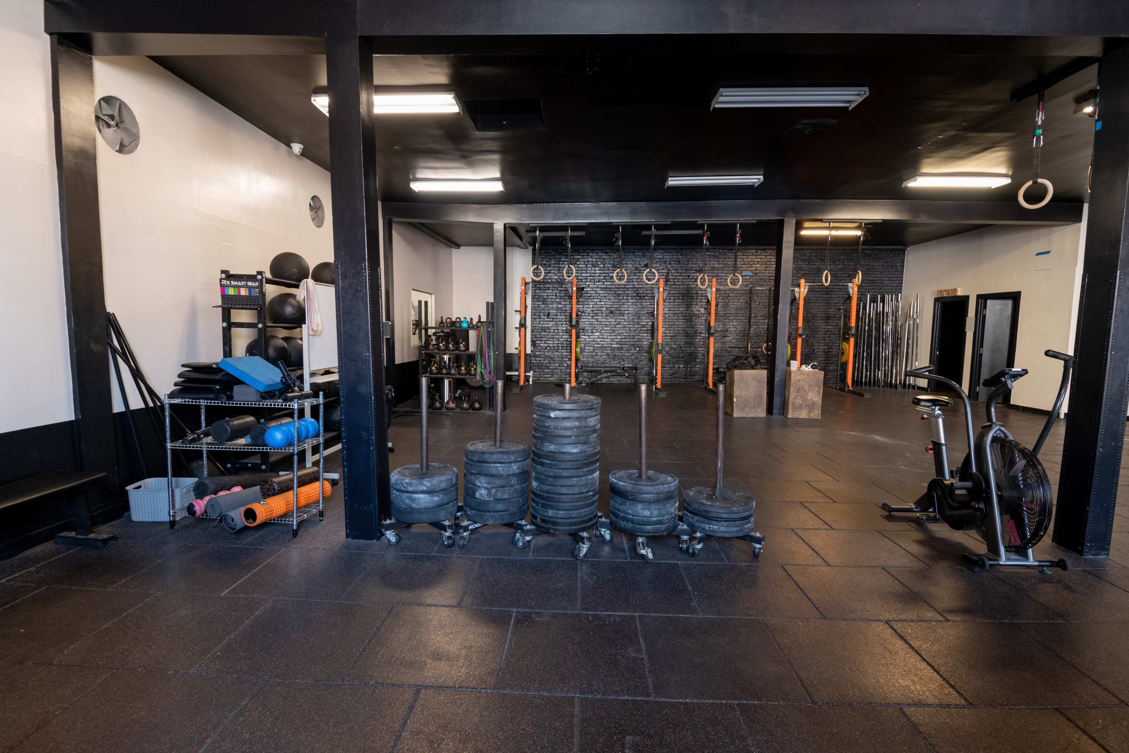 The image shows a spacious gym interior featuring weight plates stacked on the floor, exercise equipment on shelves, and rings suspended from the ceiling.