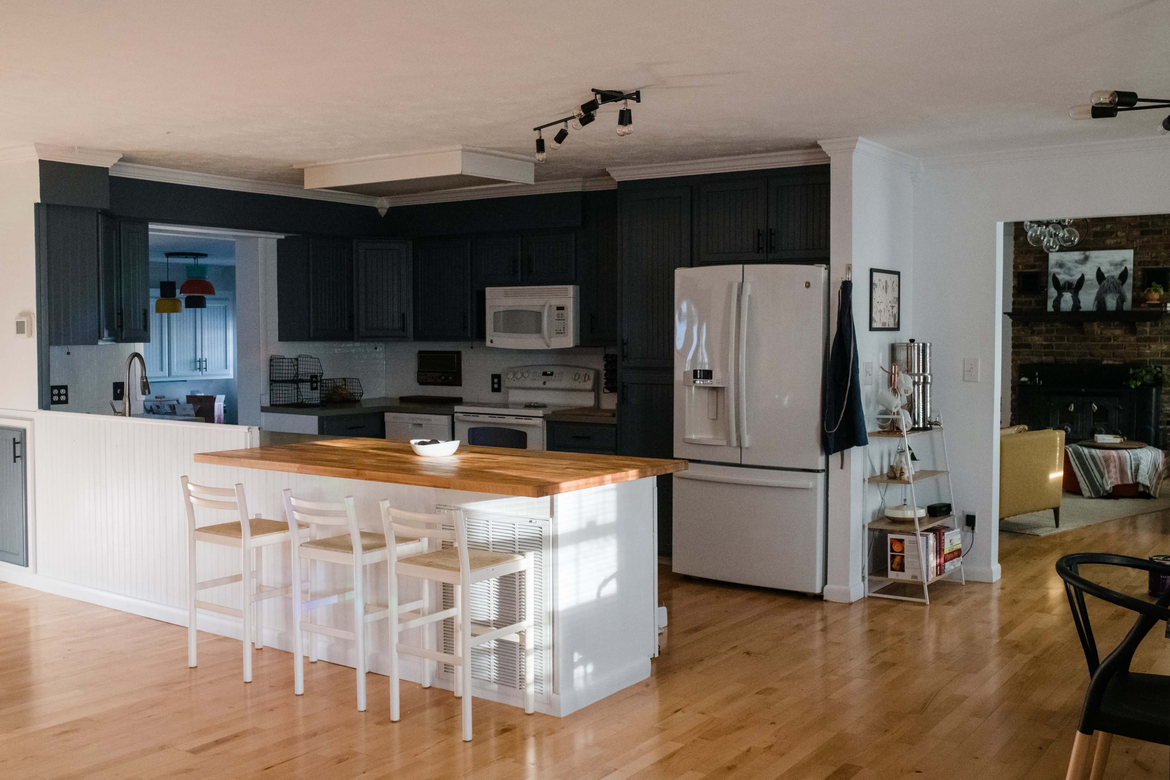 A modern kitchen features a wooden island with four white stools, surrounded by dark cabinetry and appliances, with an open layout leading to a living area.