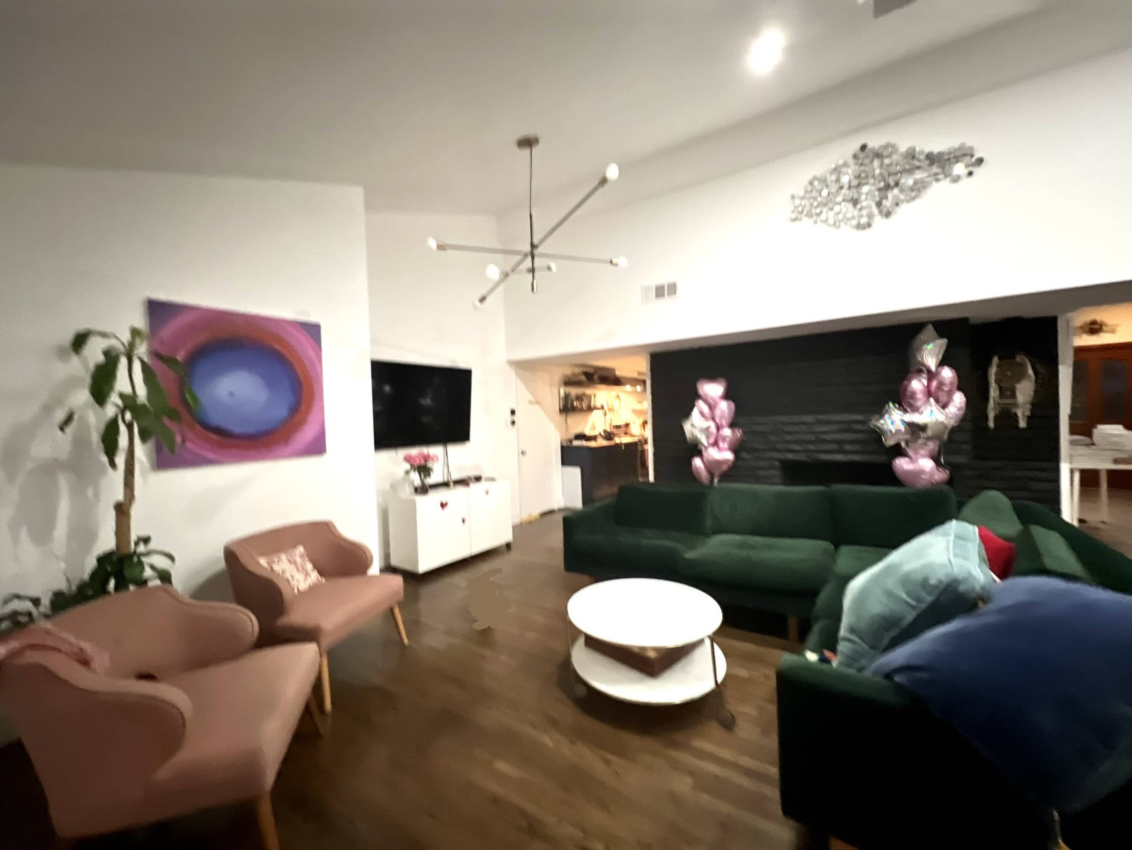 A modern living room features a green sectional sofa, a round coffee table, and pink accent chairs, with decorative elements including a large abstract painting and balloons in the corner.