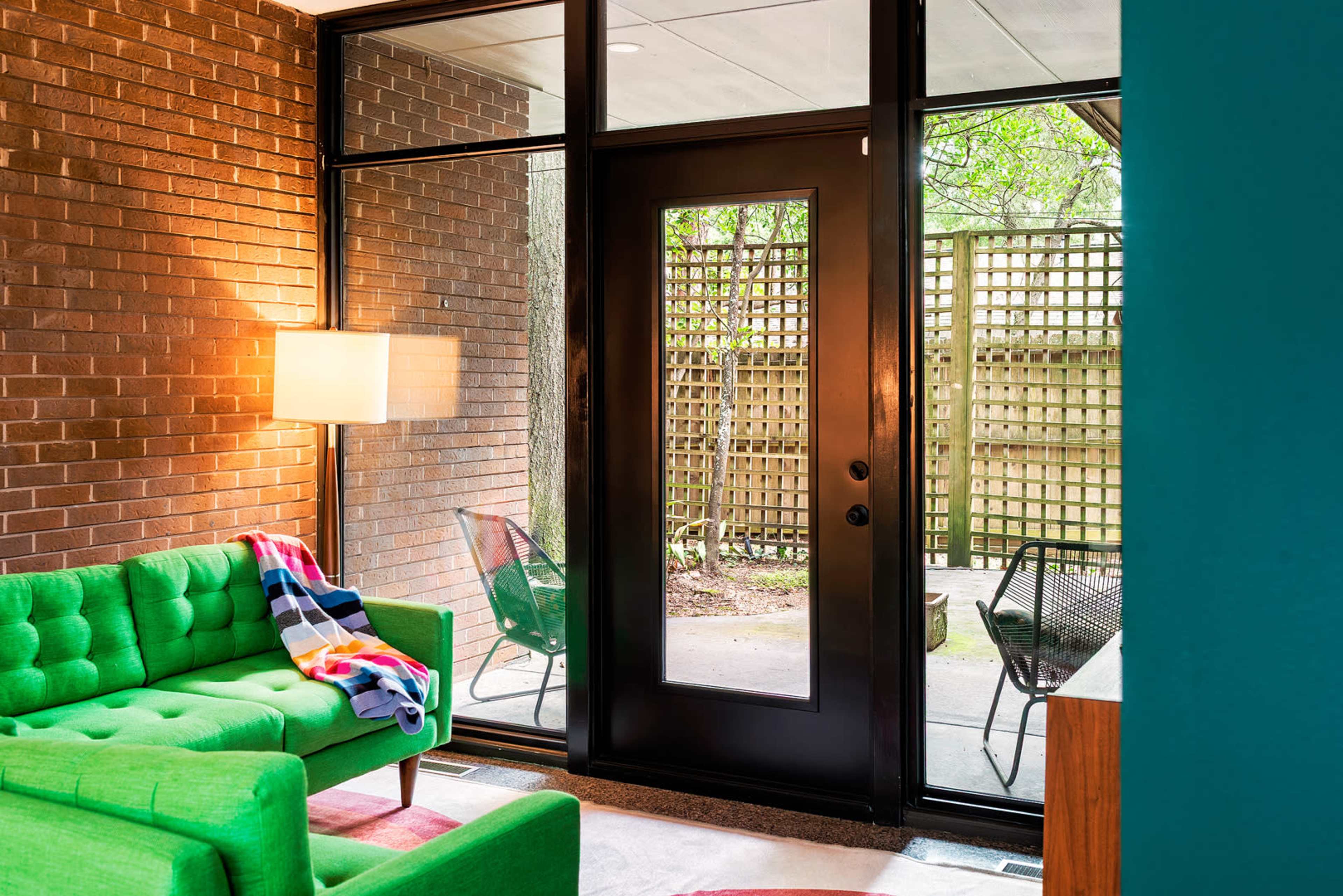 The image shows a modern living area featuring a green couch, a floor lamp, and a view of an outdoor patio through glass doors.