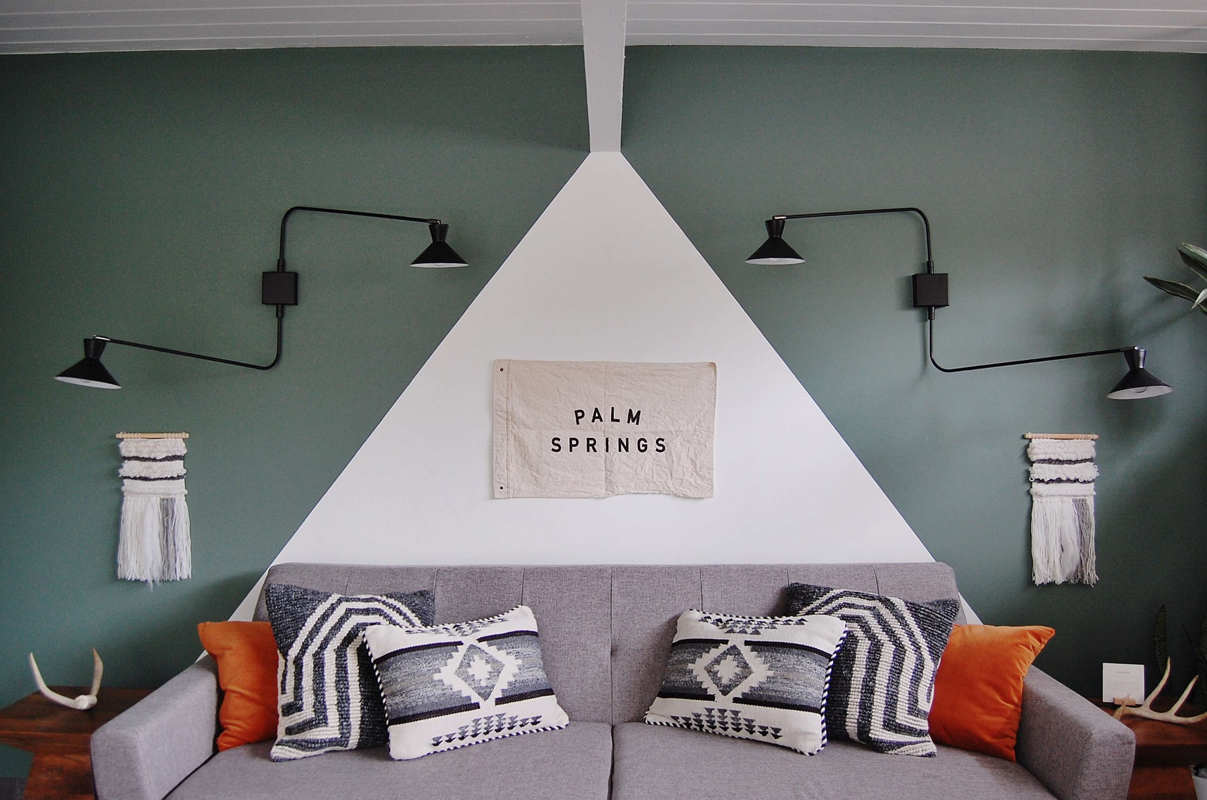 A gray sofa with patterned pillows and orange accents is positioned against a green wall featuring triangular designs and wall-mounted lights.