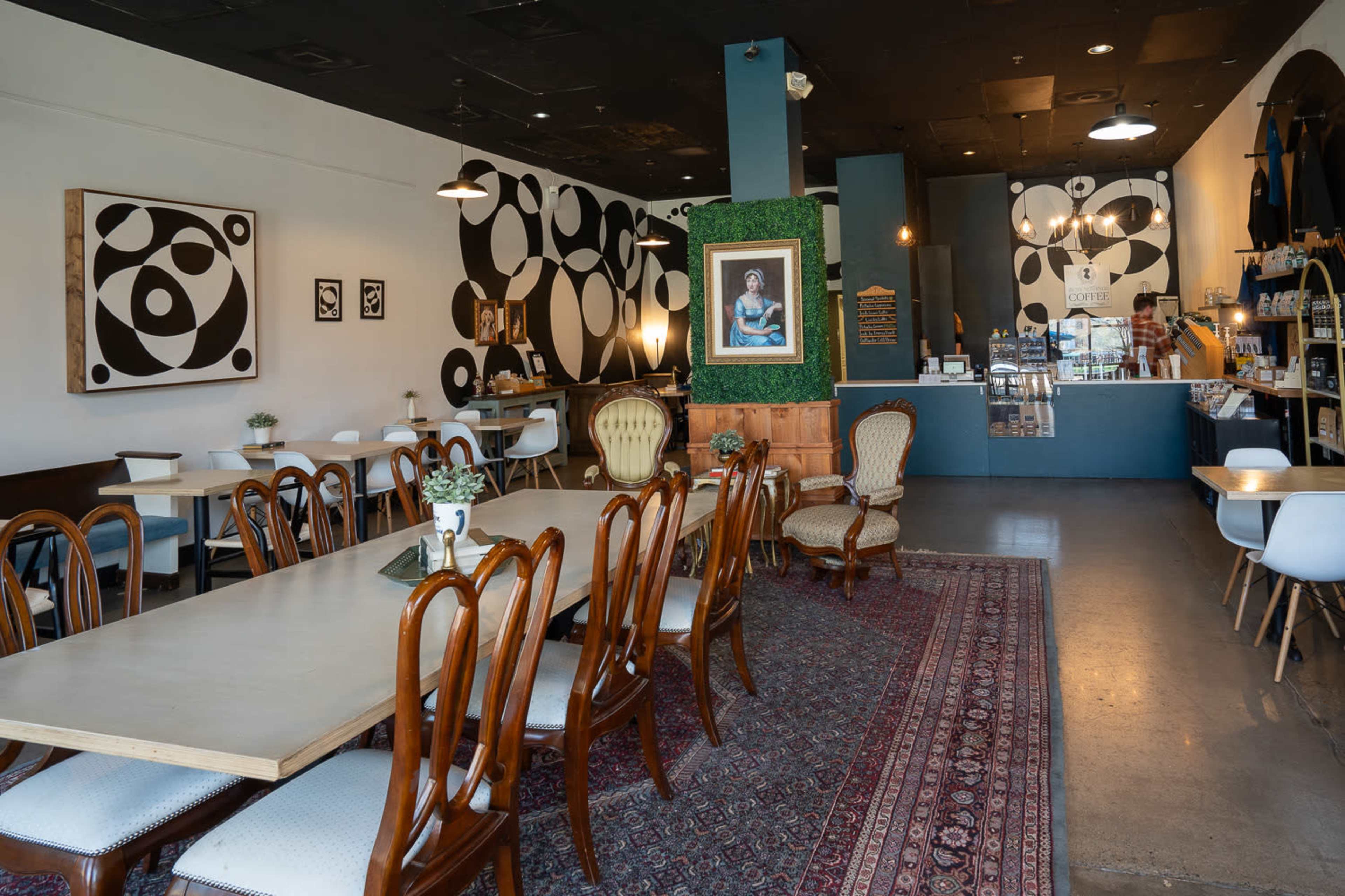 A cozy café interior with wooden tables, upholstered chairs, and a decorative rug, featuring abstract wall art and a counter displaying various food and drink items.