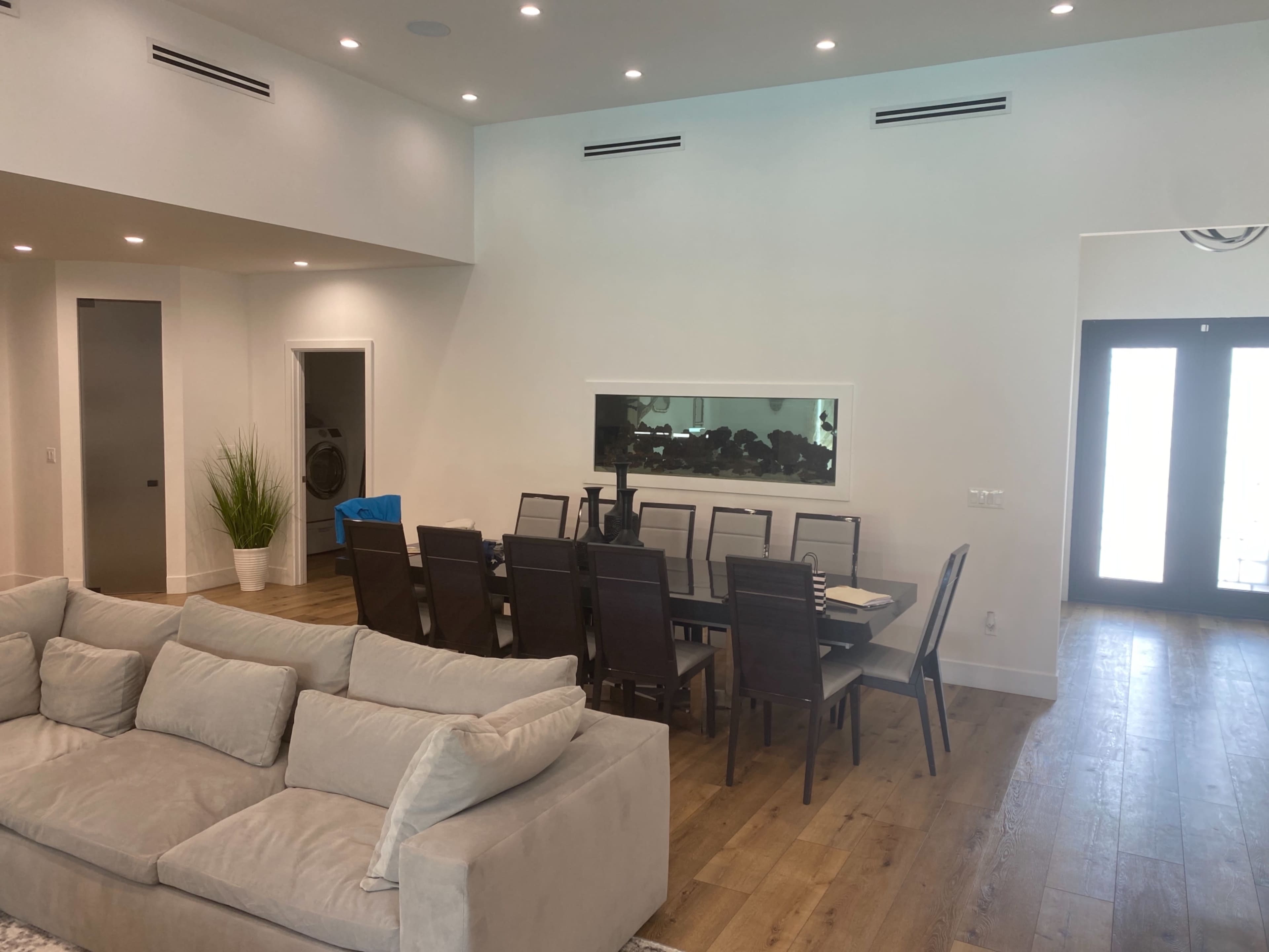 A spacious living area features a light-colored sofa and a dining table with eight chairs, adjacent to a large window and double doors leading outside.