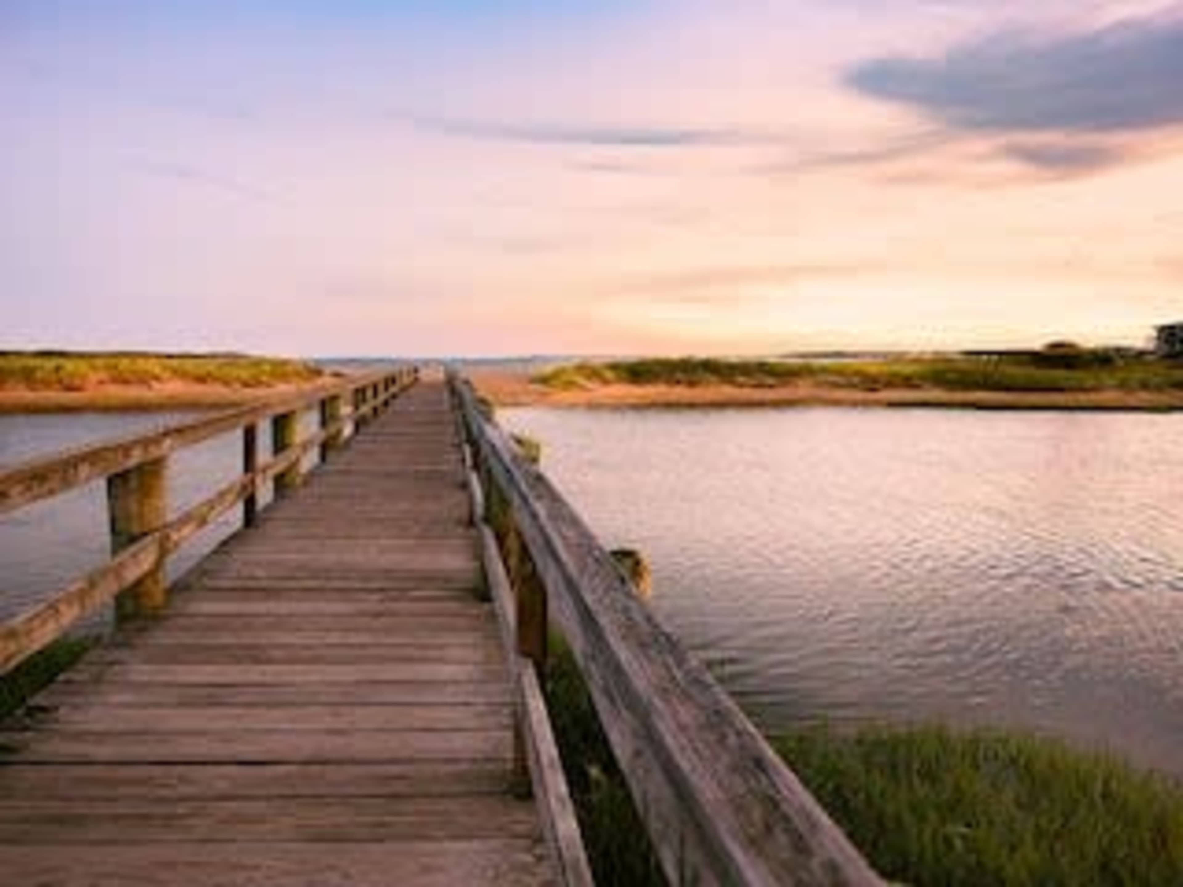 A wooden walkway extends over a calm body of water, surrounded by lush greenery and a pastel sky at dusk.
