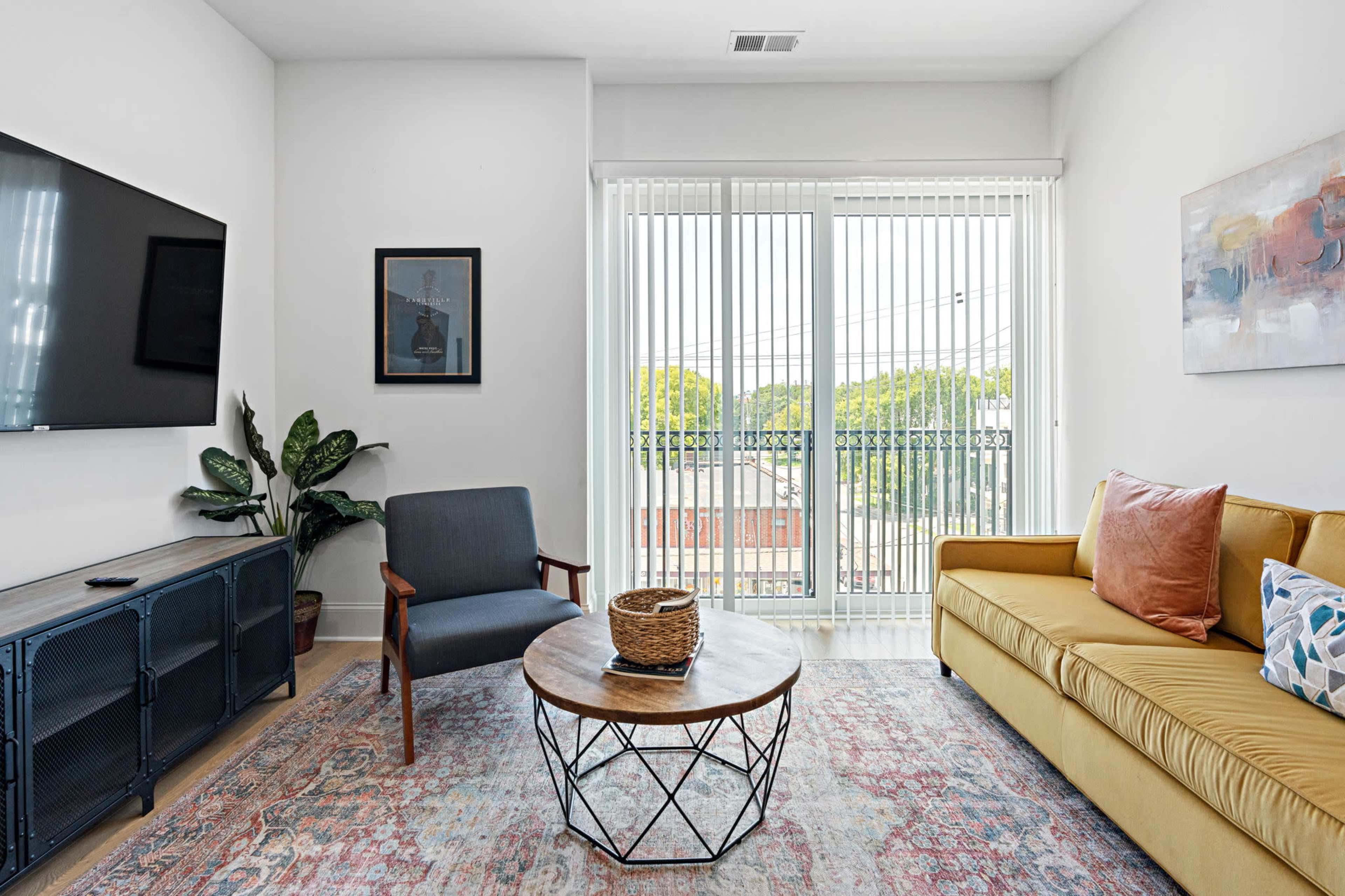 A living room features a yellow sofa, a wooden coffee table, a television on the wall, and large windows with sheer blinds.