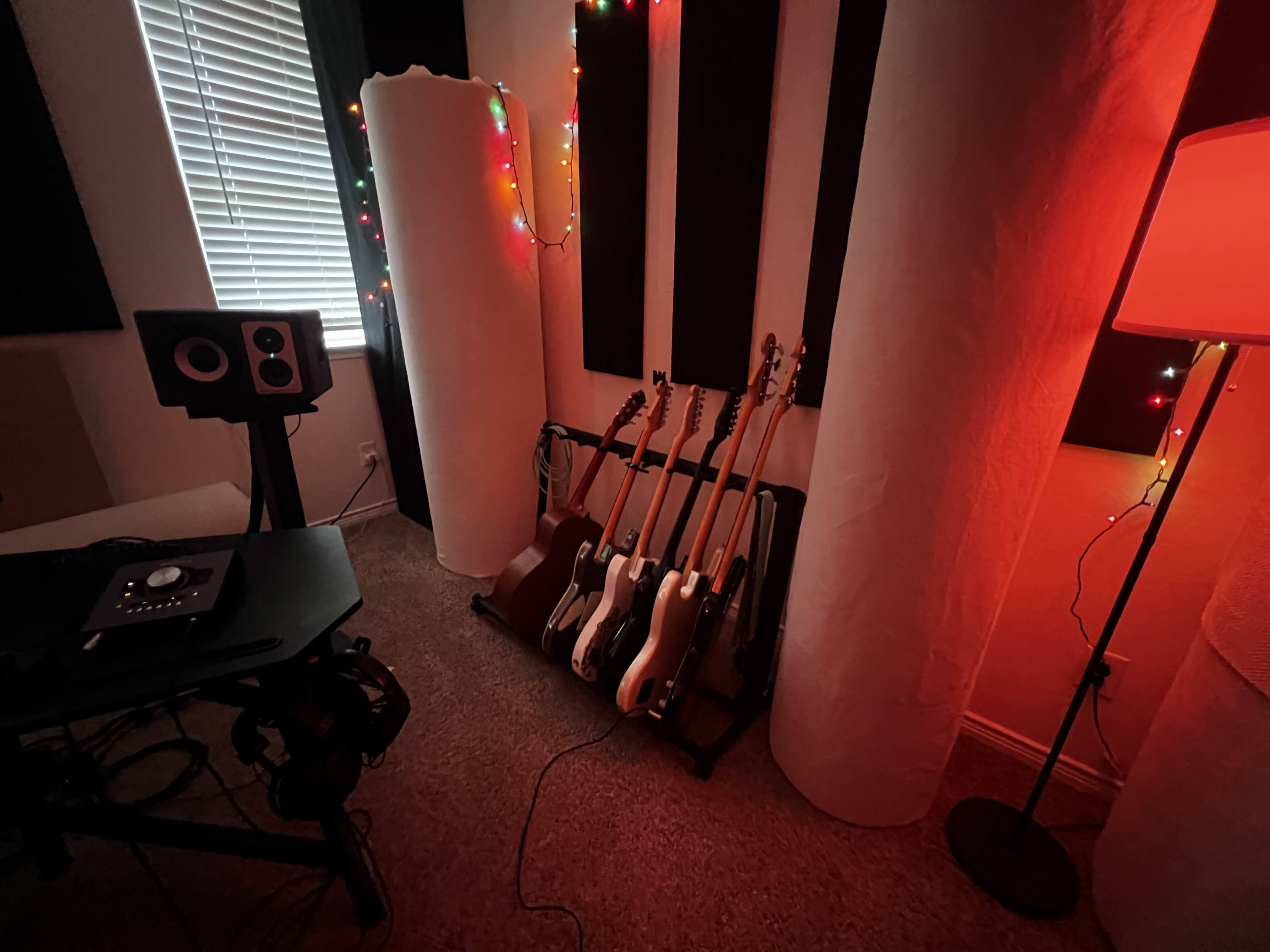 The image shows a music studio corner with several electric guitars resting on a stand next to soundproofing panels and a chair beside a computer setup.