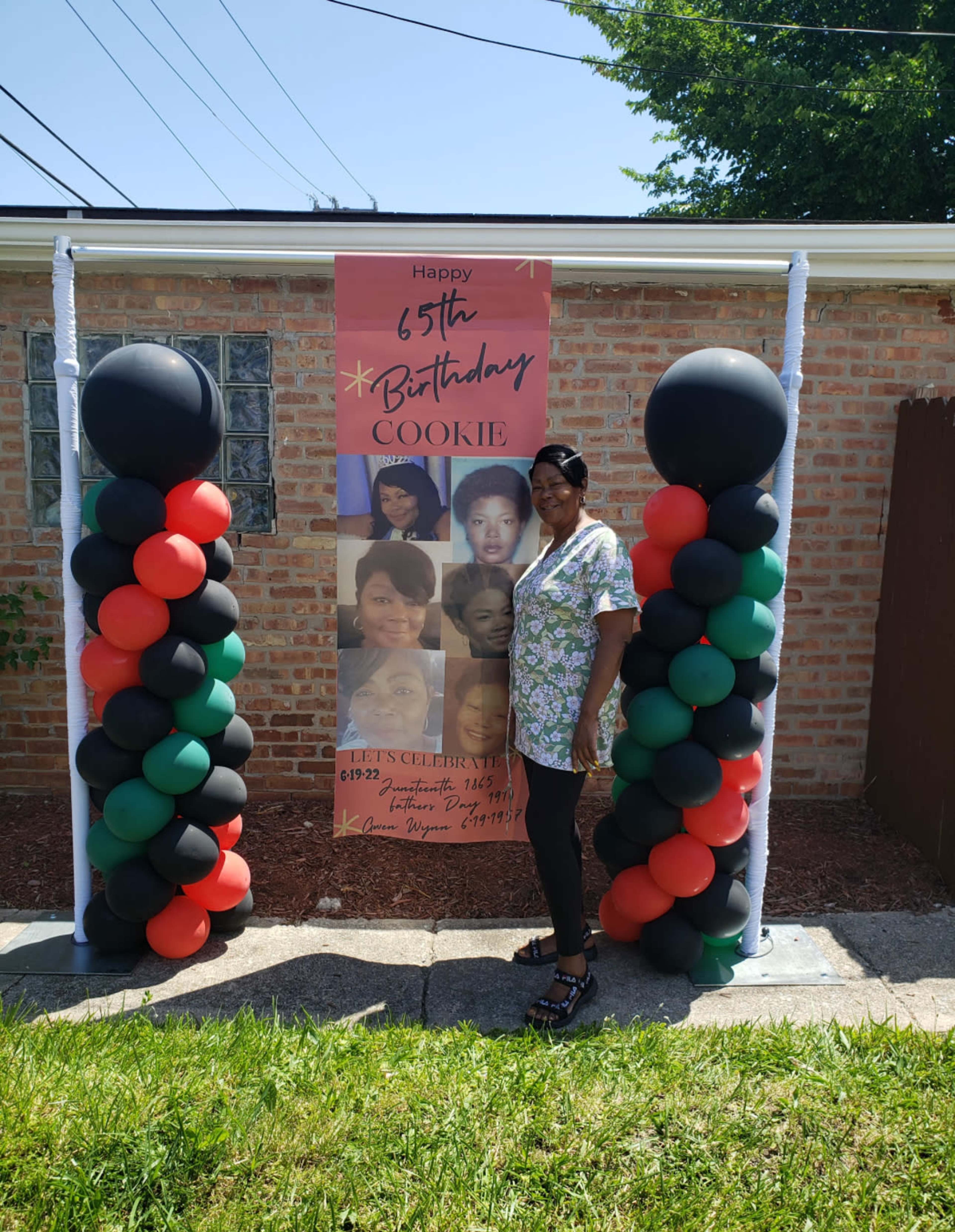 A woman stands next to a birthday banner celebrating a 65th birthday, flanked by columns of black, red, and green balloons.