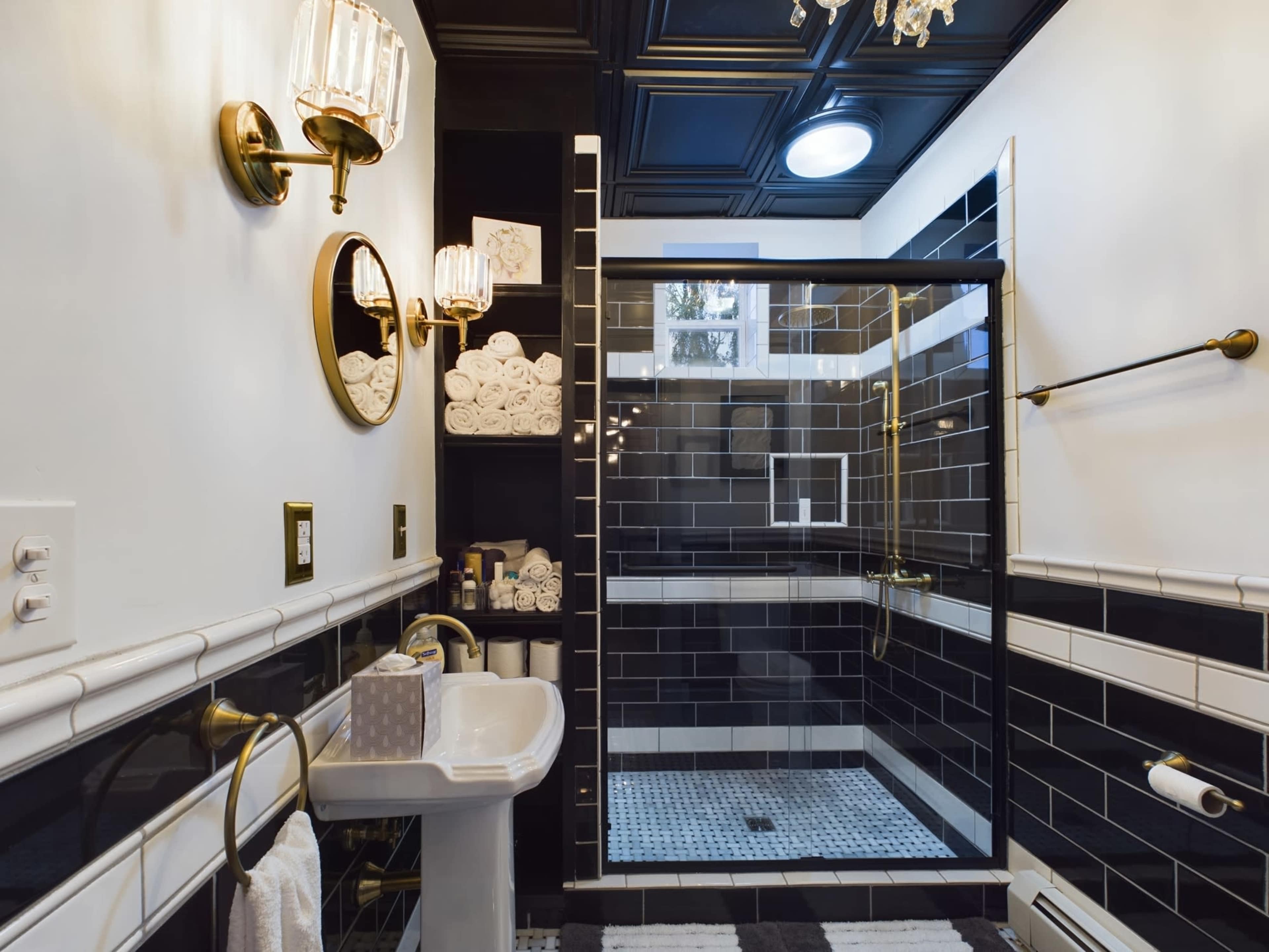 The bathroom features a glass shower enclosure, black tiled walls, a white pedestal sink, and stacked white towels in decorative niches.