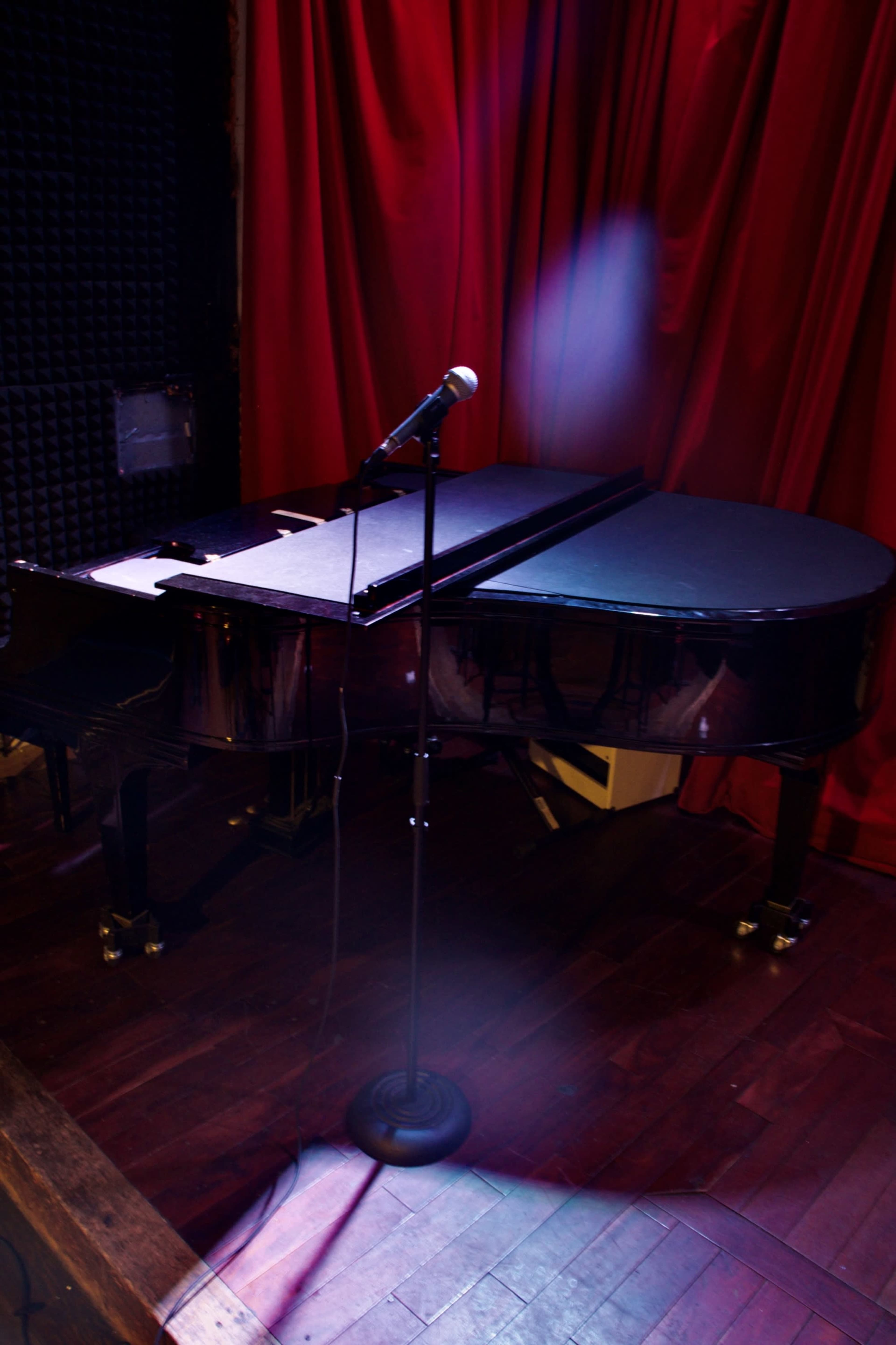A black grand piano is positioned next to a microphone stand in front of a red curtain.