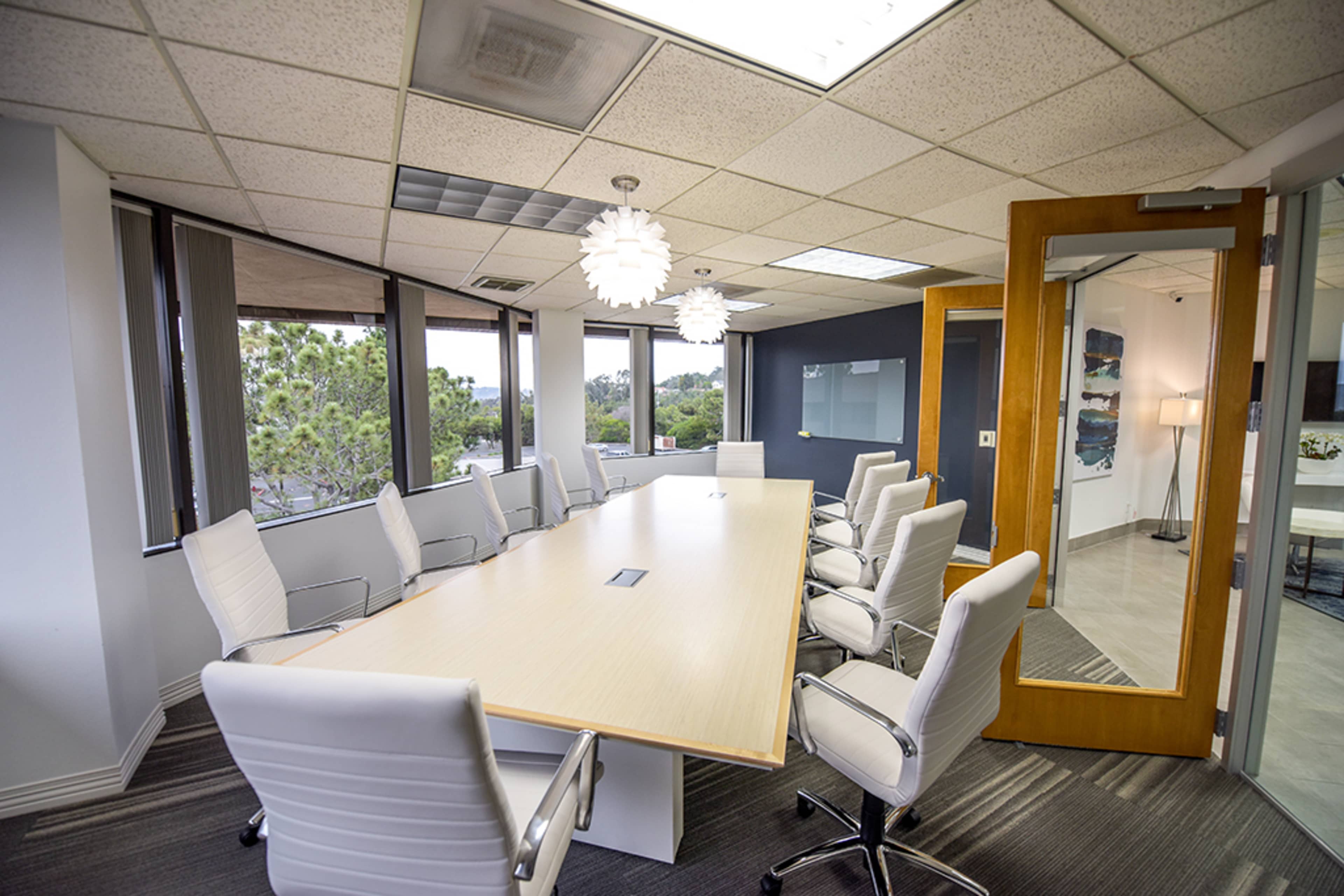 A modern conference room features a long wooden table surrounded by white chairs and large windows allowing natural light.
