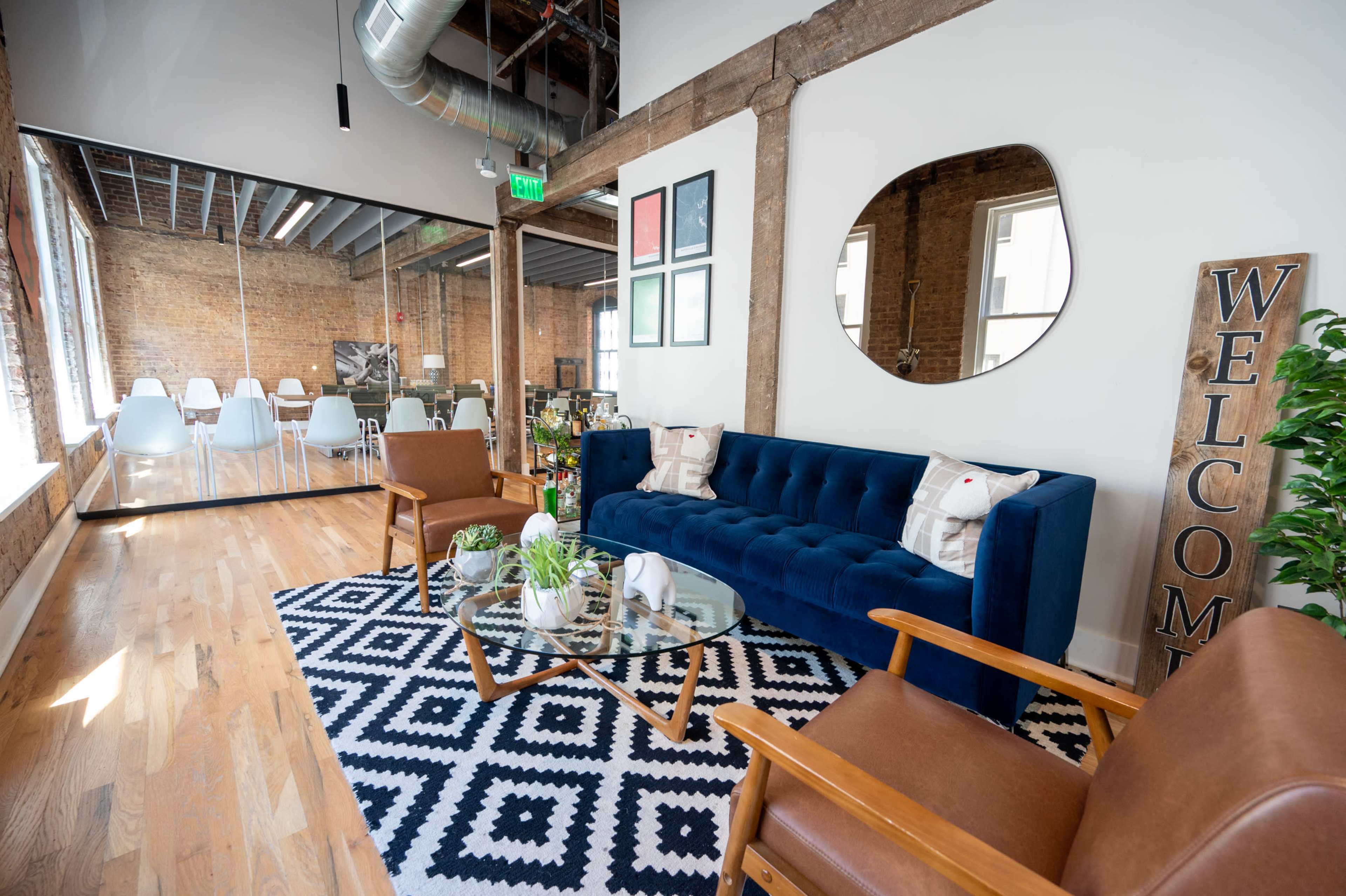 A bright lounge area features a blue sofa, two wooden chairs, a glass coffee table on a patterned rug, and a large mirror on the wall, with a welcoming sign nearby.