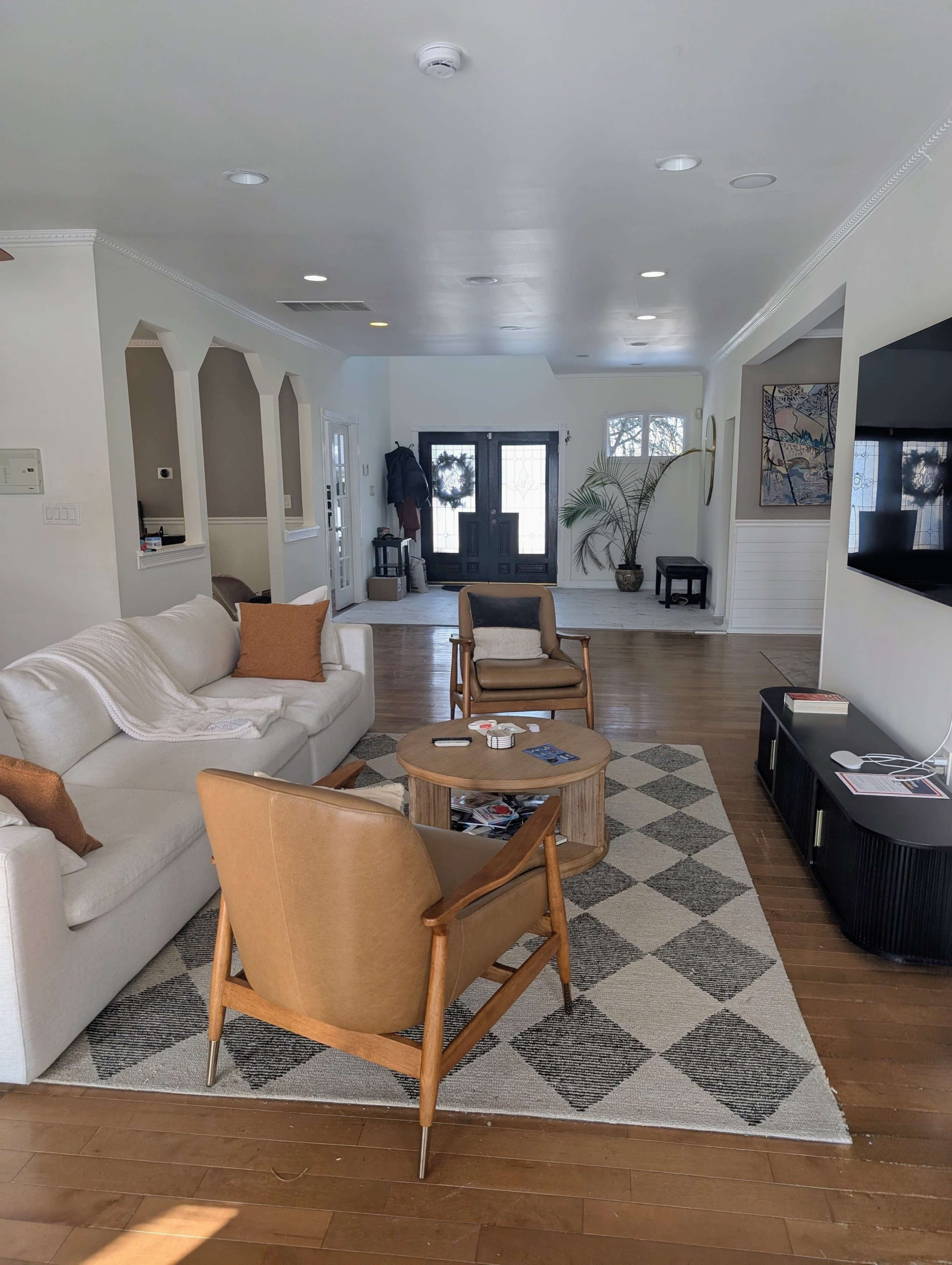 The living room features a sofa, two armchairs, a round coffee table, and a television mounted on the wall, with a neutral color palette and wooden flooring.