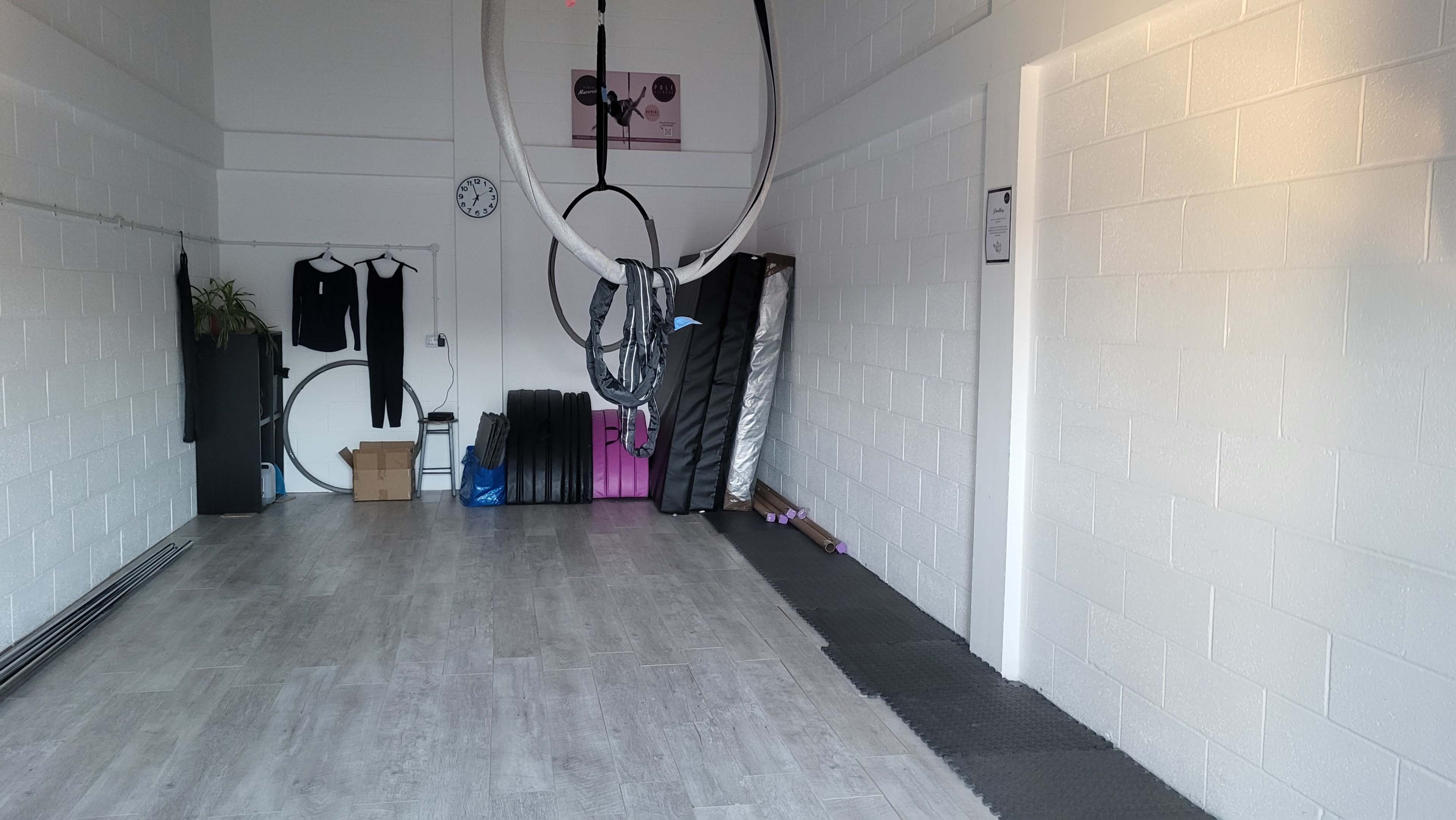 The image shows a spacious, empty studio with a wall-mounted clock, aerial hoops hanging from the ceiling, and various workout mats stacked along one wall.