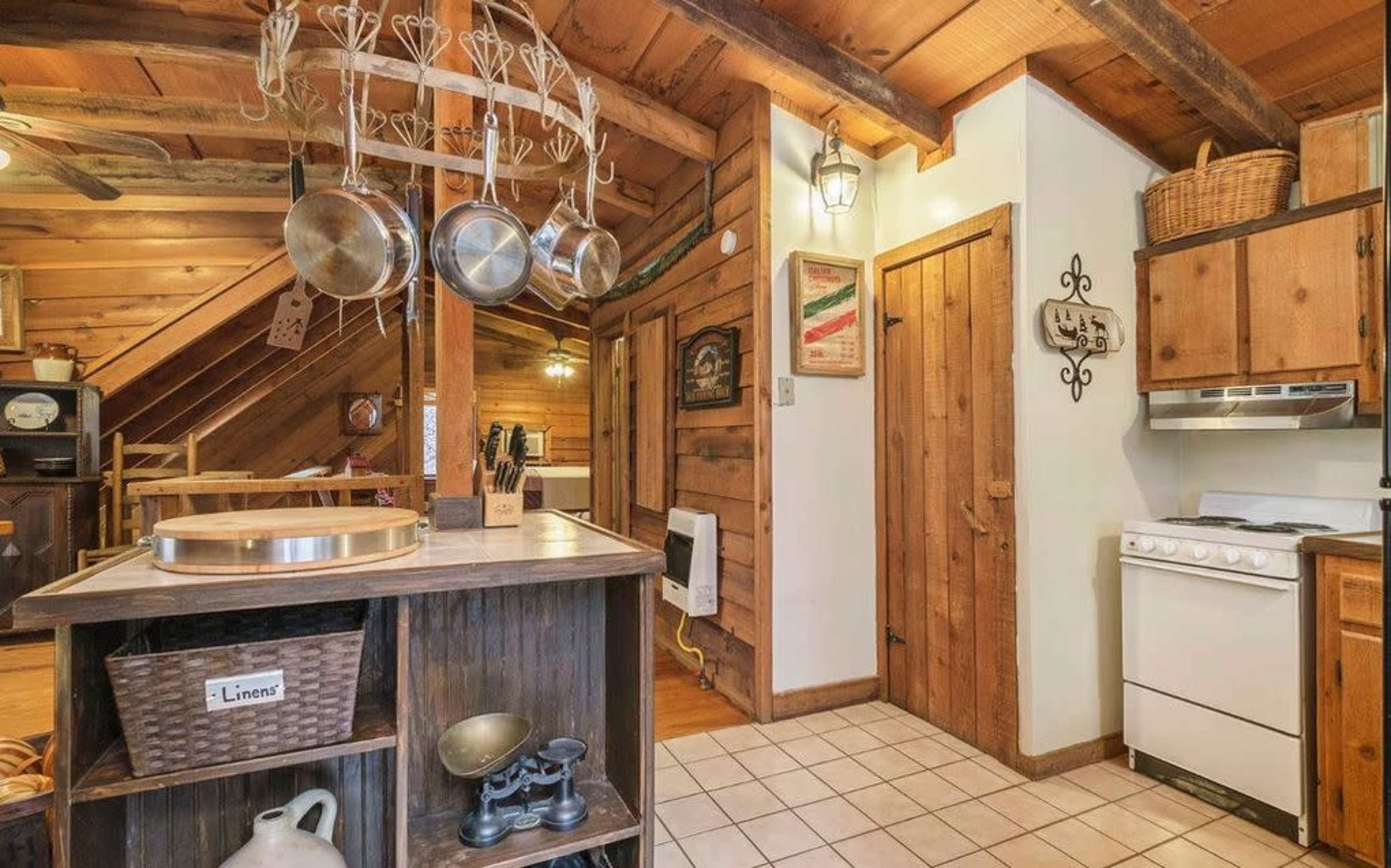 A rustic kitchen with wooden beams, a stove, and hanging pots, connected to a small dining area.