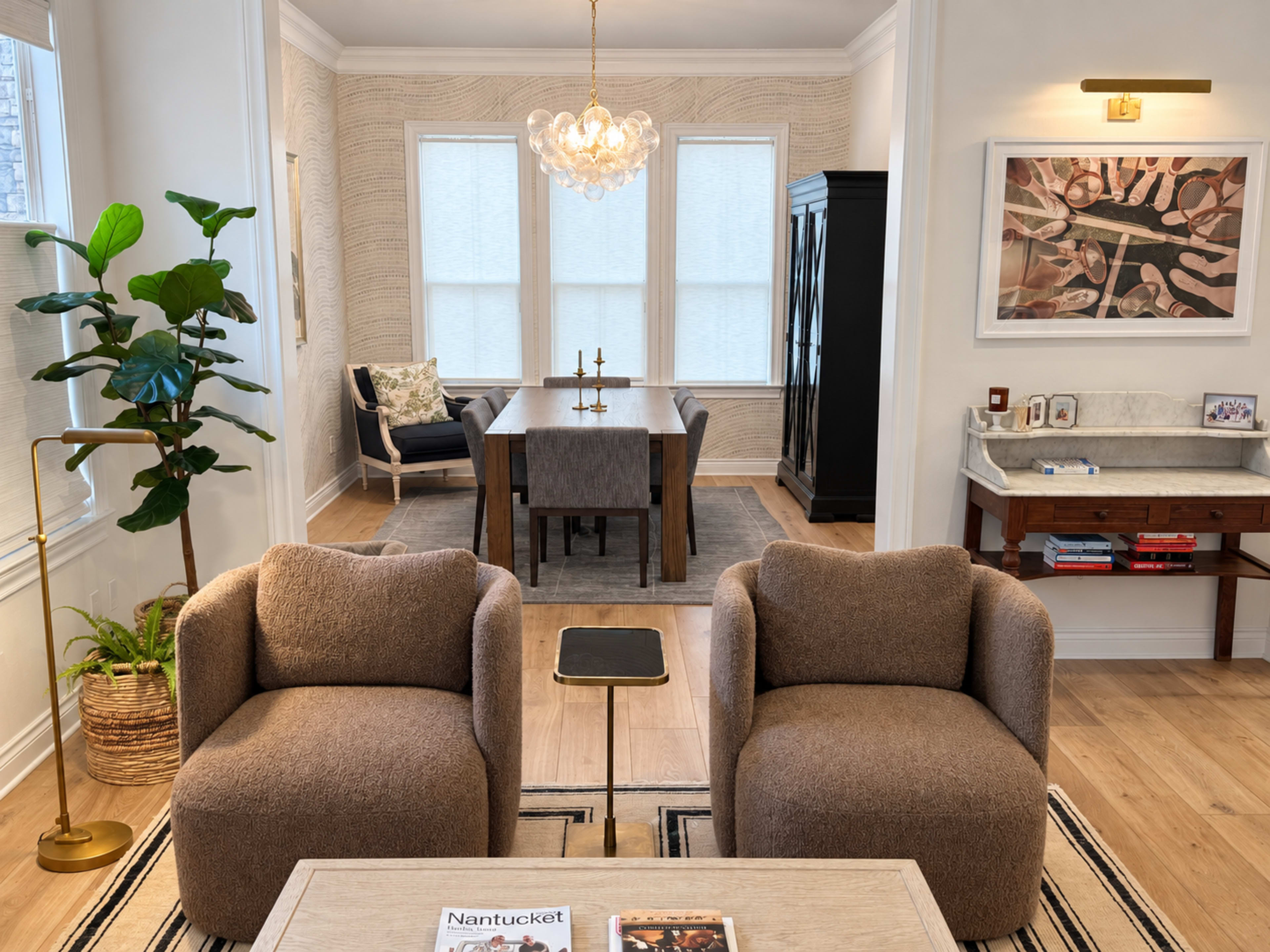 The scene features a modern living area with two soft armchairs facing a coffee table, while a dining area with a large table and a decorative cabinet is visible in the background.