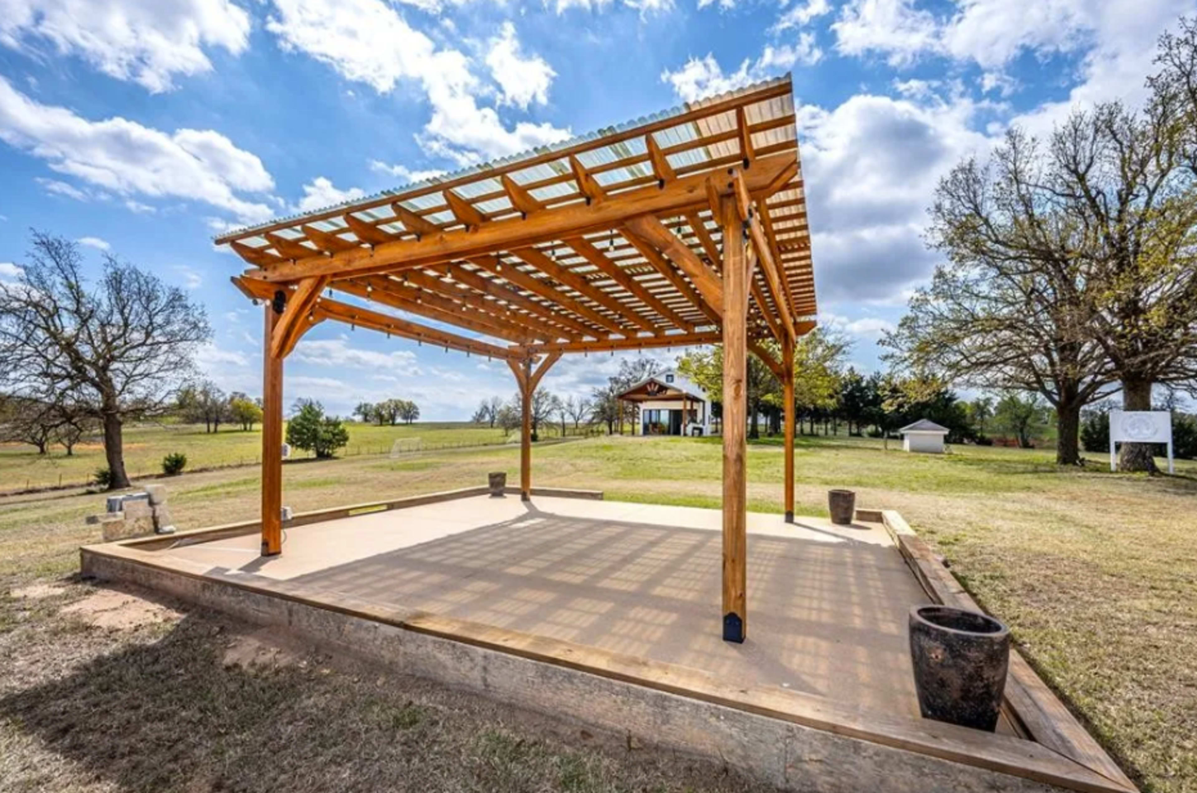 The image shows a wooden pergola on a concrete slab in an open field with trees and a building in the background.