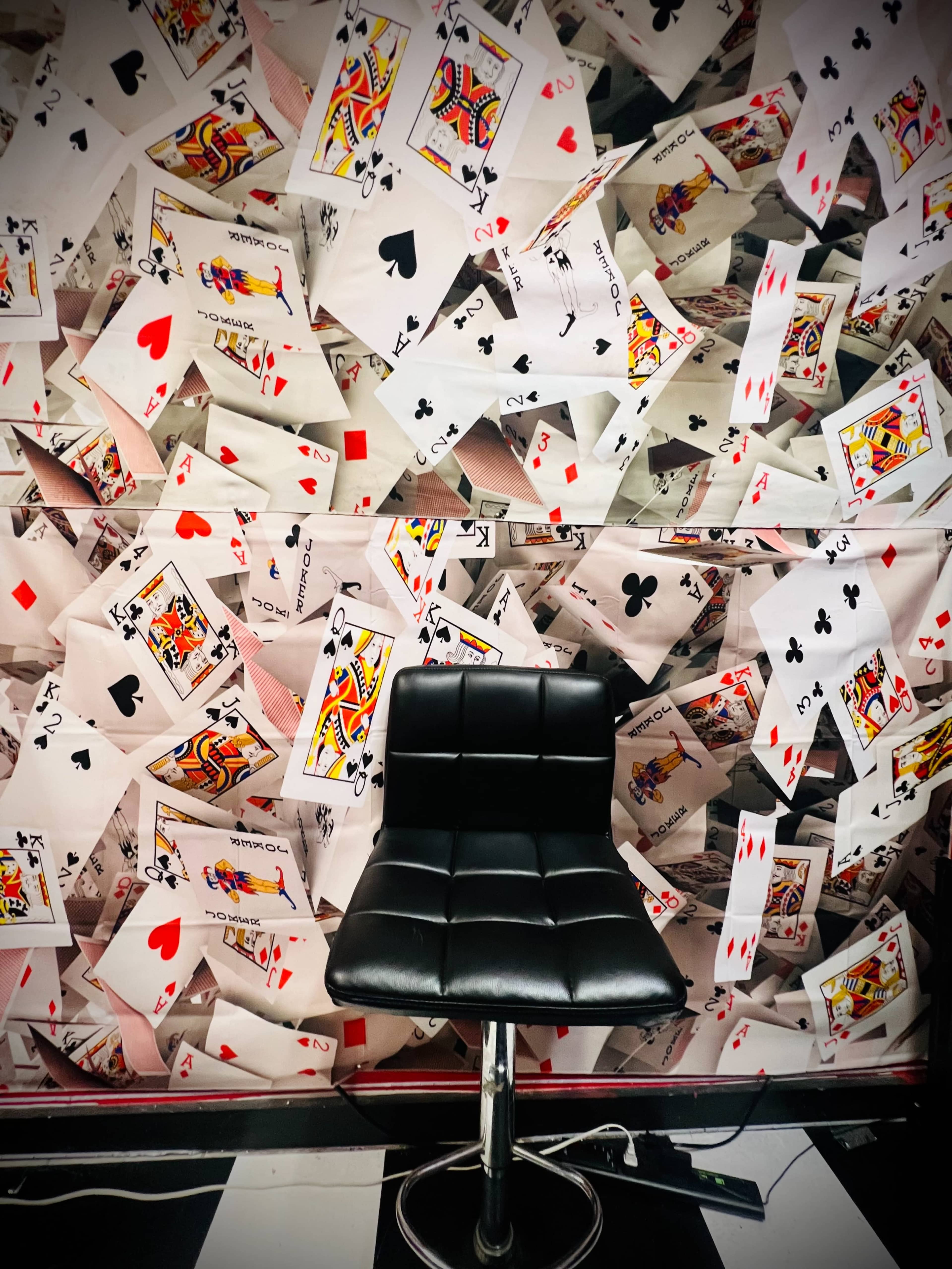 A black bar stool is positioned in front of a wall covered with scattered playing cards.