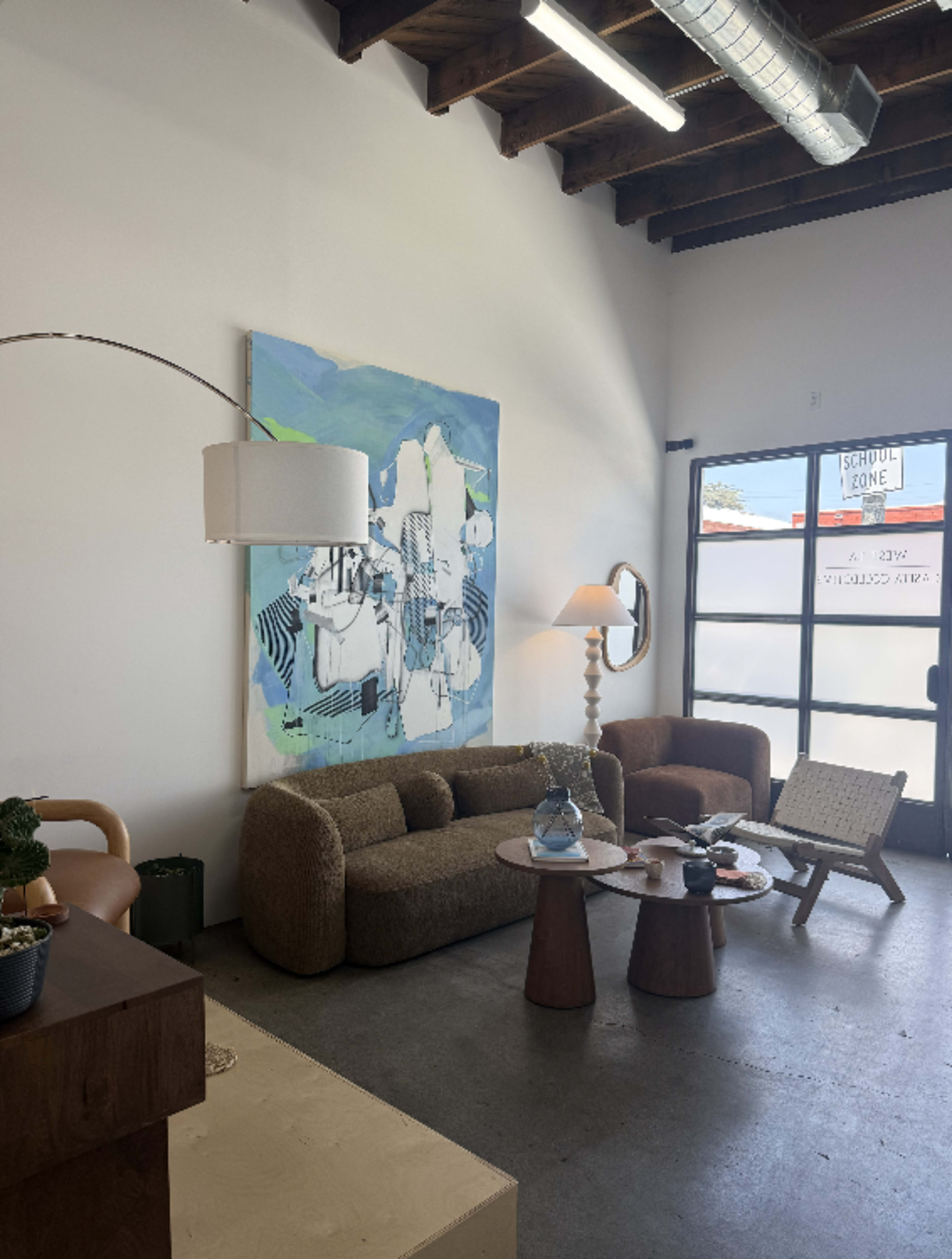 The image shows a modern interior of a living space featuring a brown sofa, a chair, a coffee table, and a large abstract painting on the wall.
