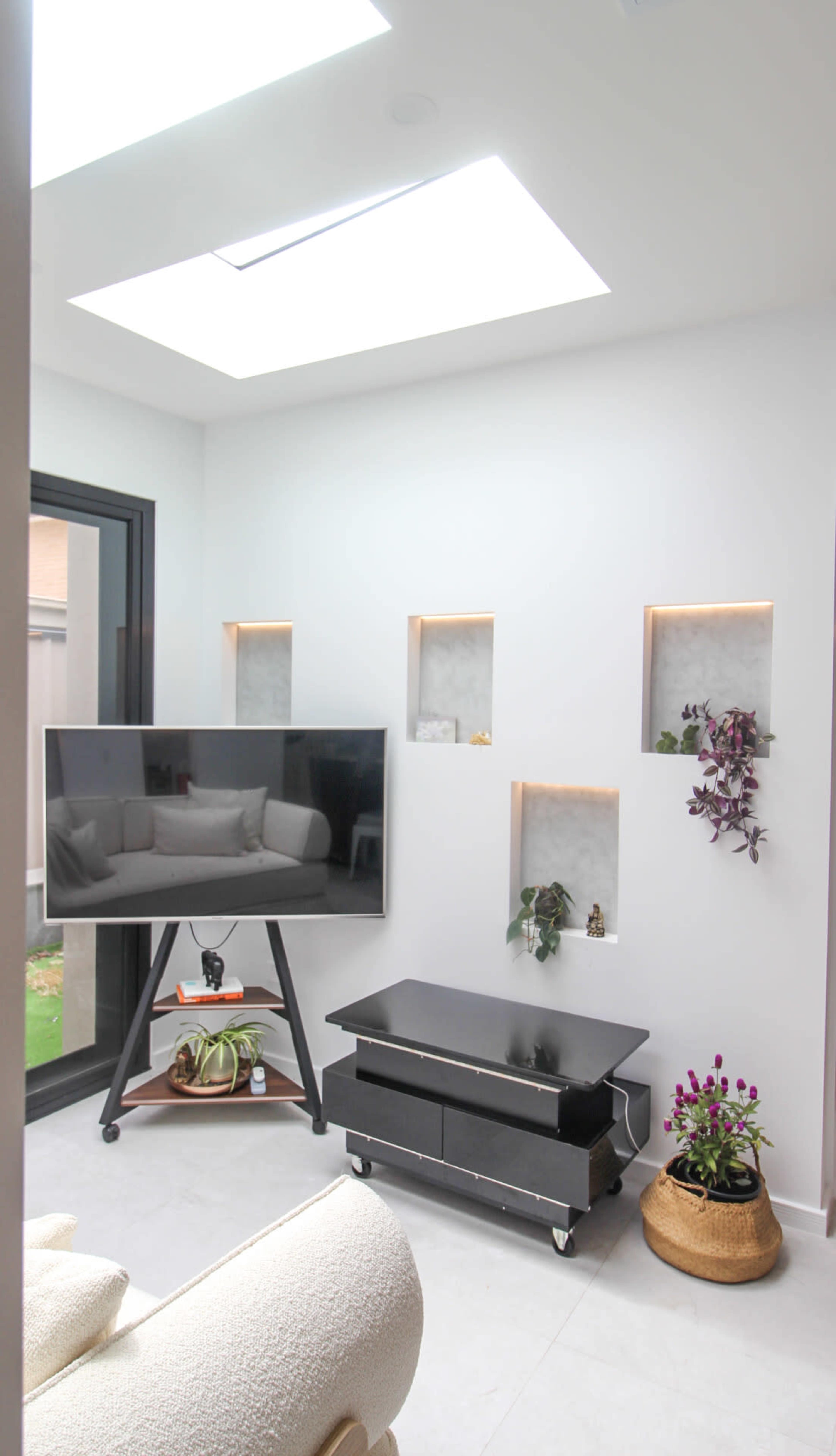 The image shows a modern sunlit room featuring a large flat-screen TV on a stand, a low black coffee table, and wall niches with subtle lighting and decorative plants.