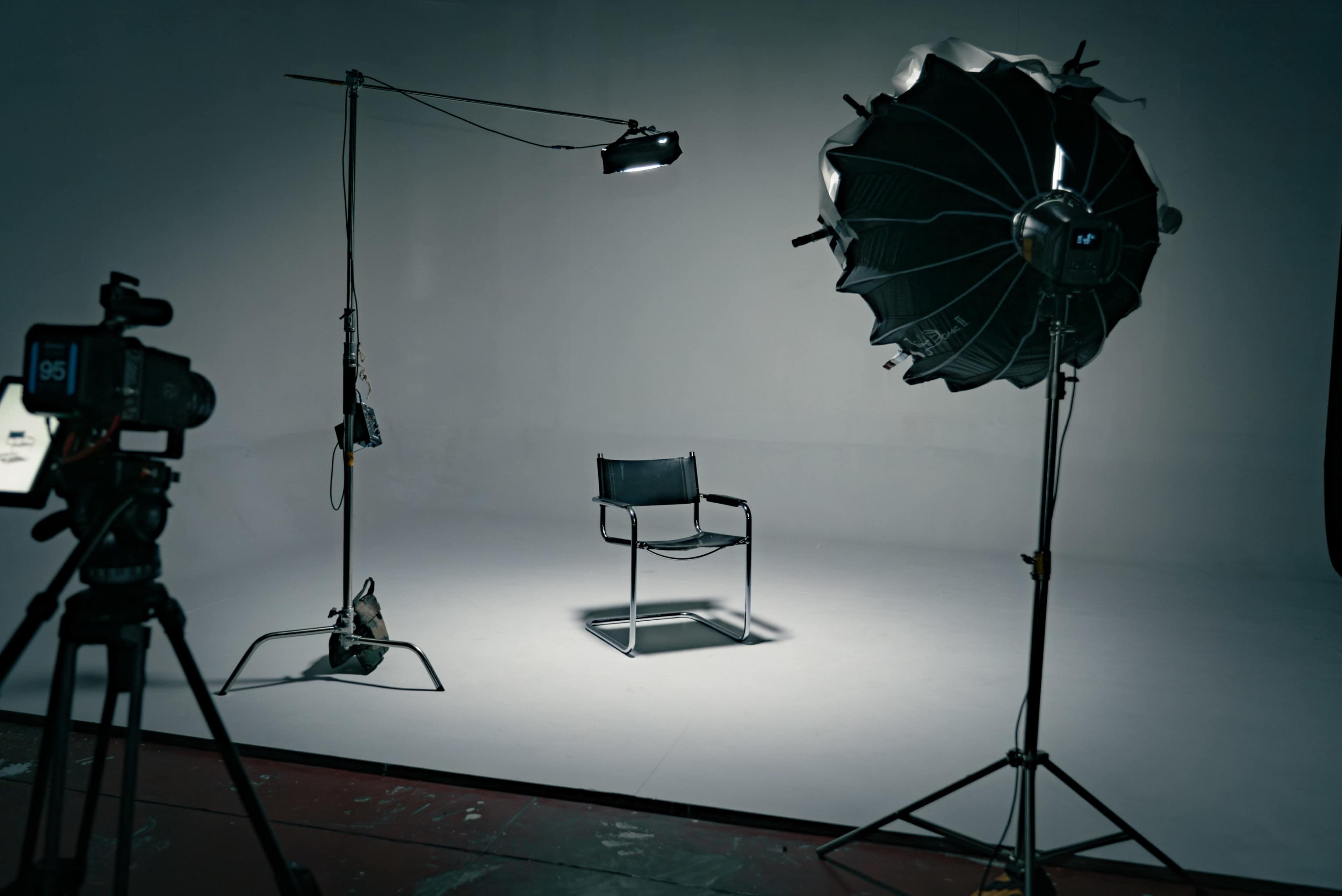 A photography studio setup includes a spotlight, a light umbrella, a camera on a tripod, and an empty chair in front of a plain backdrop.
