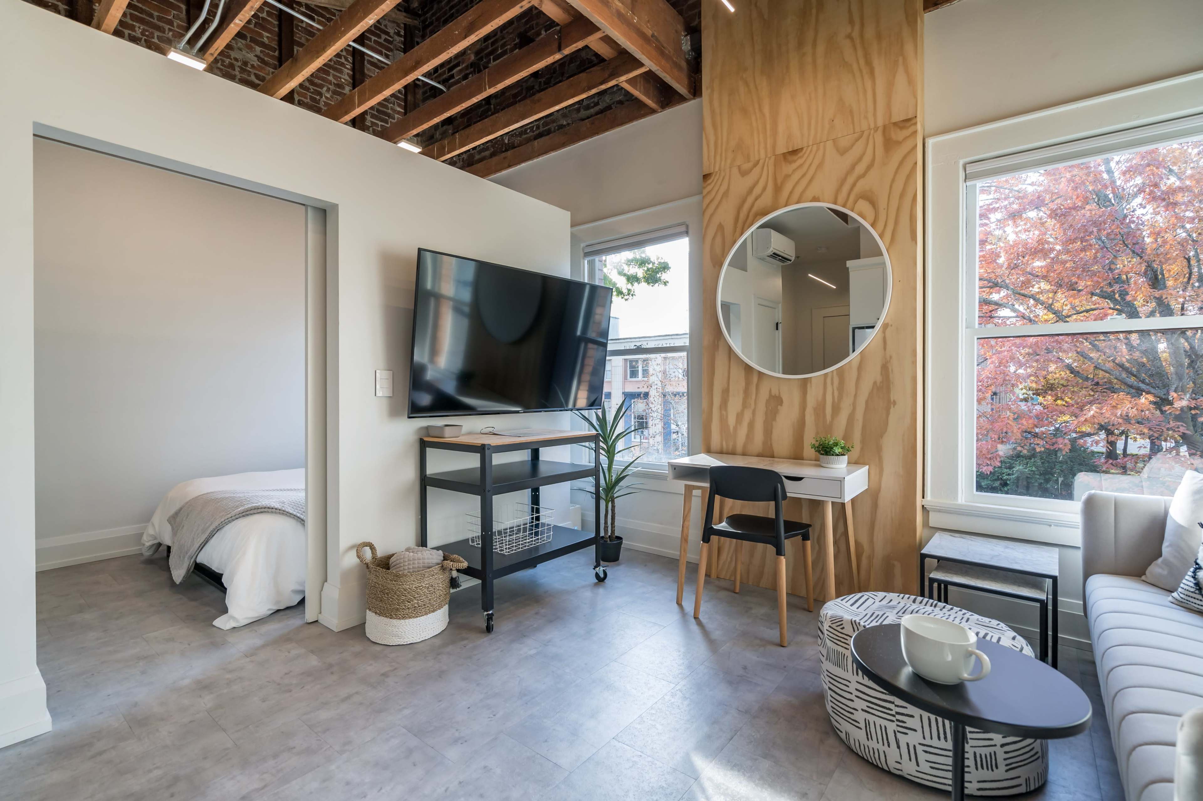 A modern studio apartment featuring a television mounted on a wooden wall, a round mirror, a small desk and chair, and a view of an outdoor autumn scene through a window.