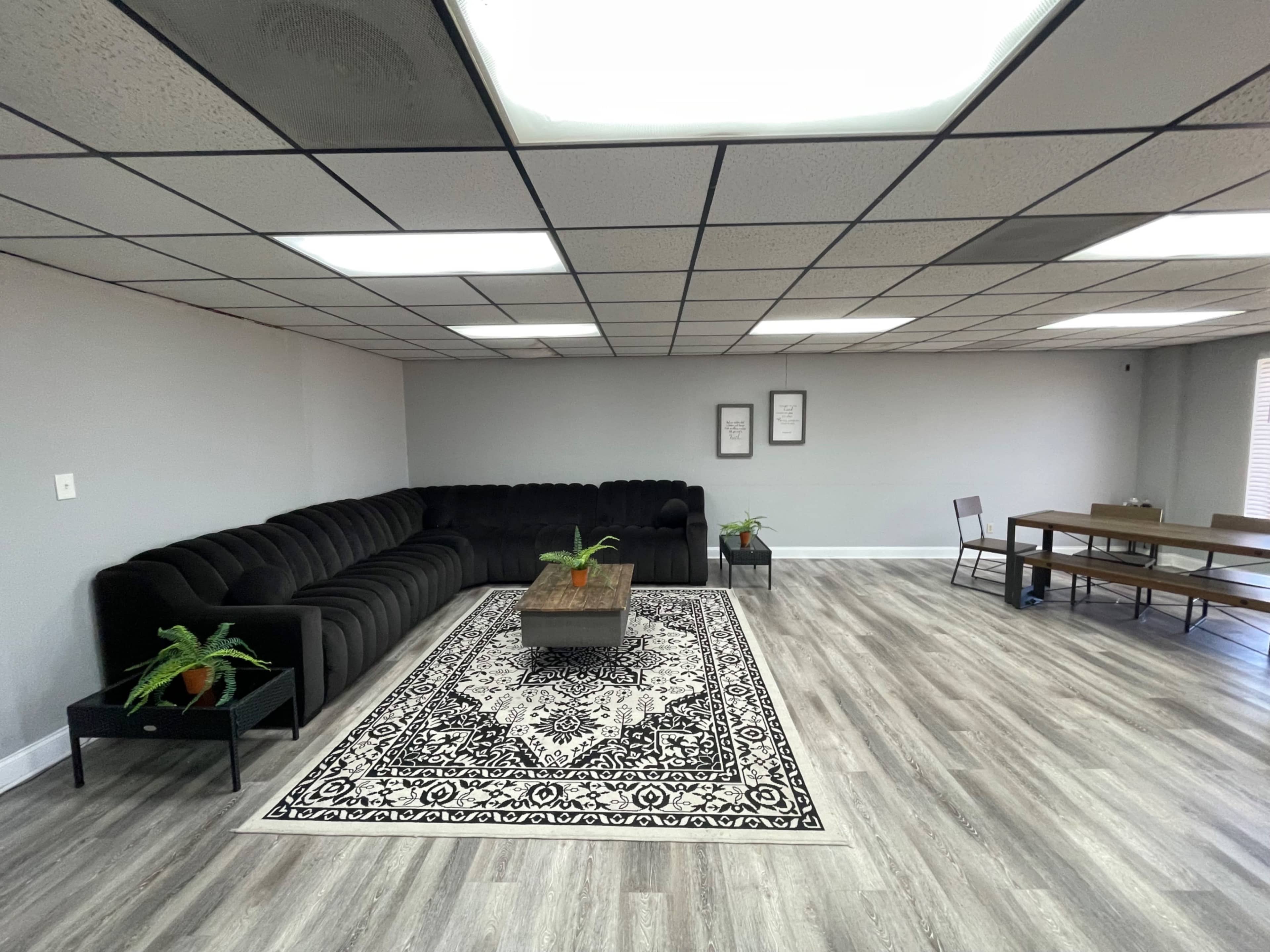 The image shows a spacious, modern room with a dark sectional sofa, a patterned area rug, small potted plants, and a wooden dining table with chairs.