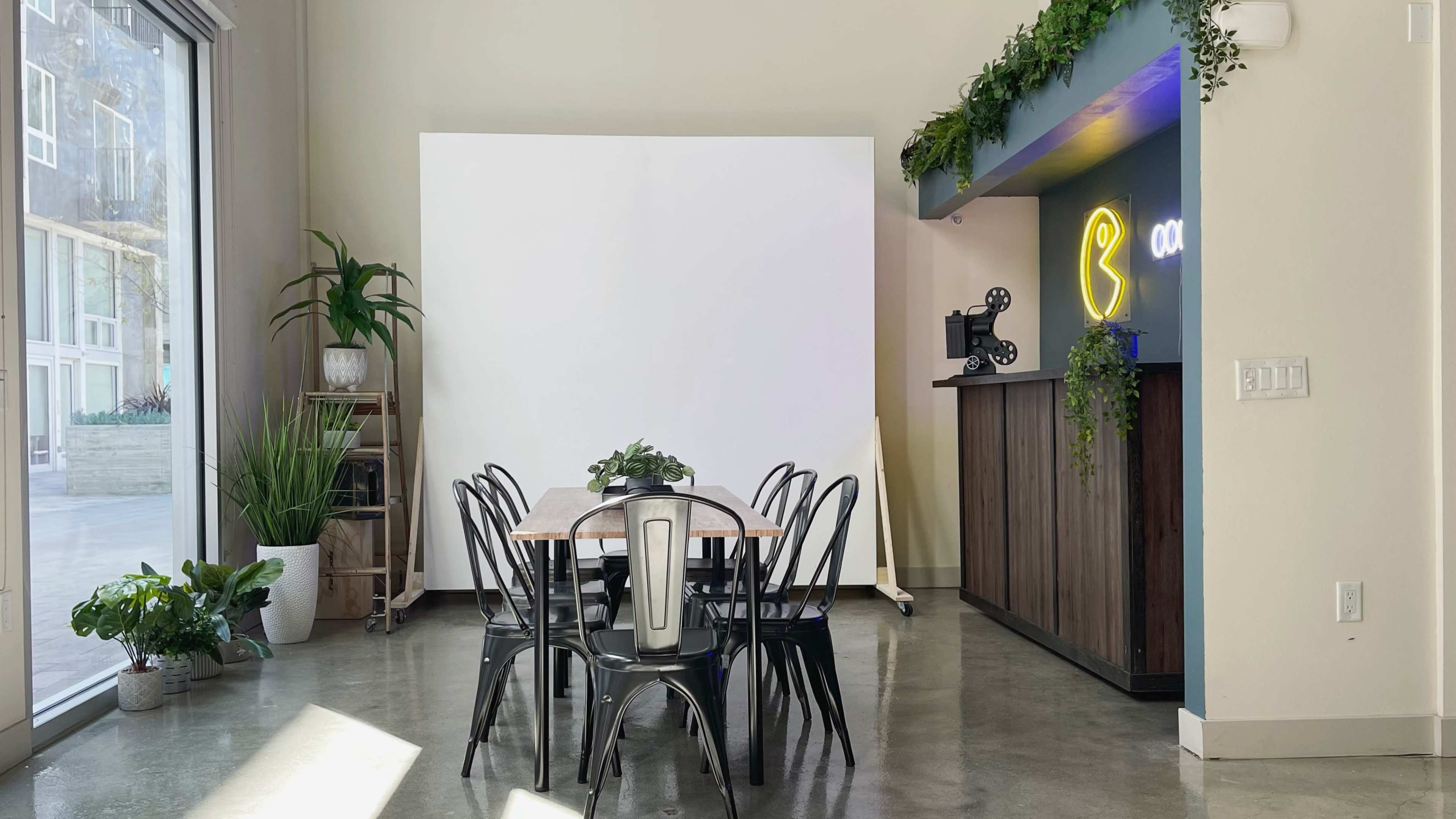 The image shows a modern interior space featuring a dining table surrounded by black chairs, a large blank white wall, and a bar area with neon lights and plants.