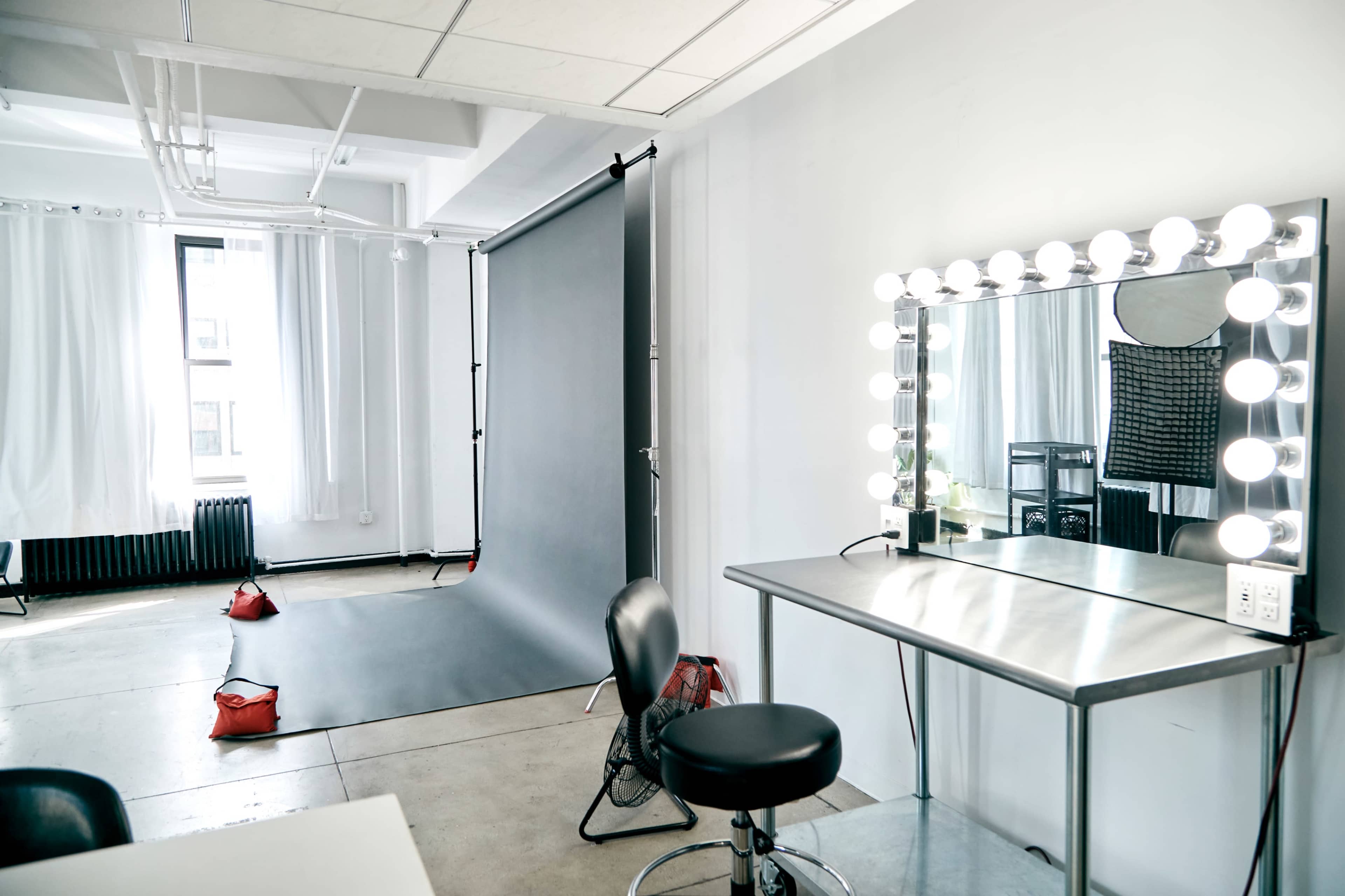 A photography studio featuring a black backdrop, a vanity mirror with lights, and a stool beside a metal table.