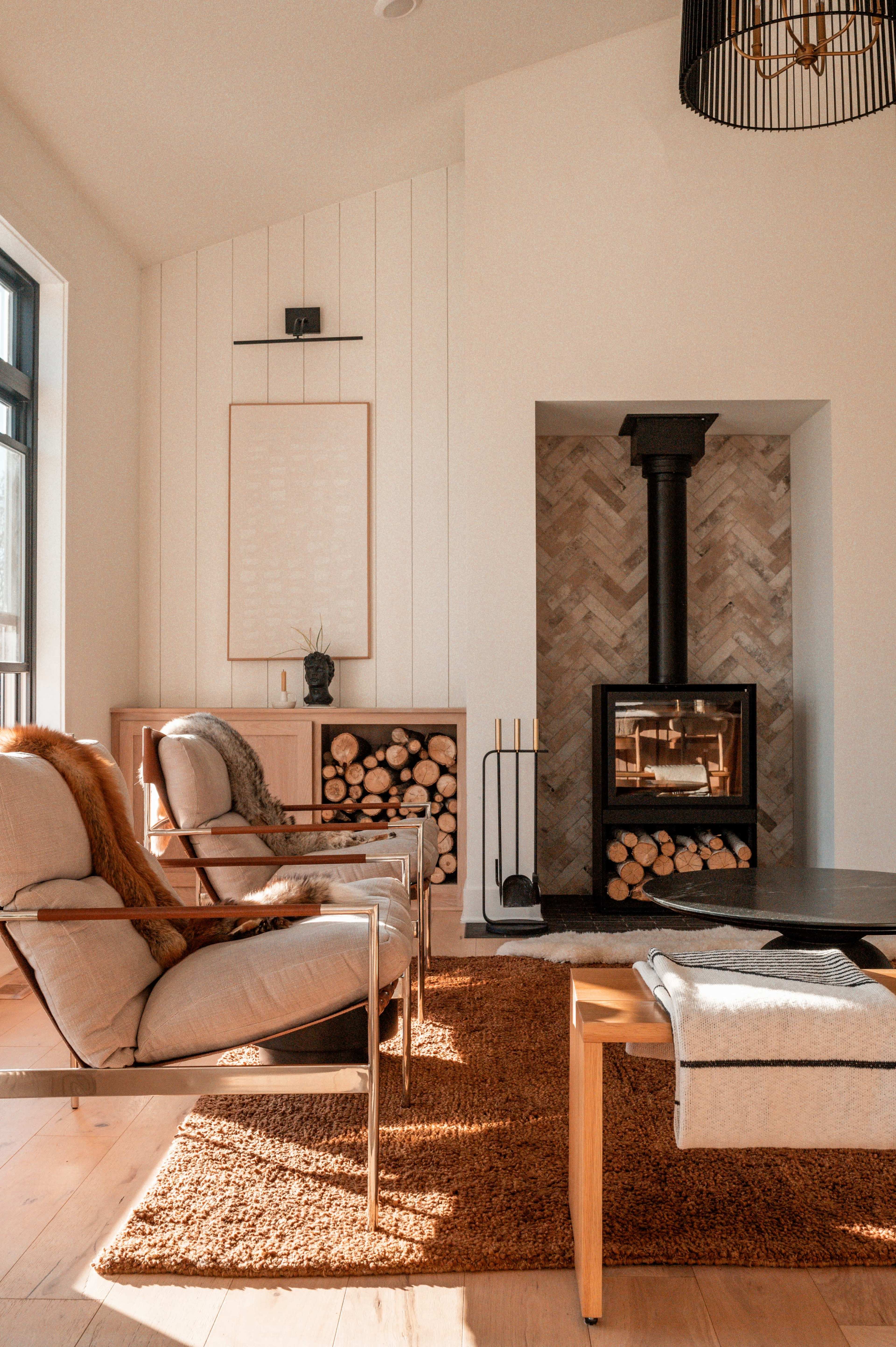 The image shows a modern living room featuring two armchairs, a coffee table, and a fireplace surrounded by stacked firewood and herringbone-patterned wall tiles.