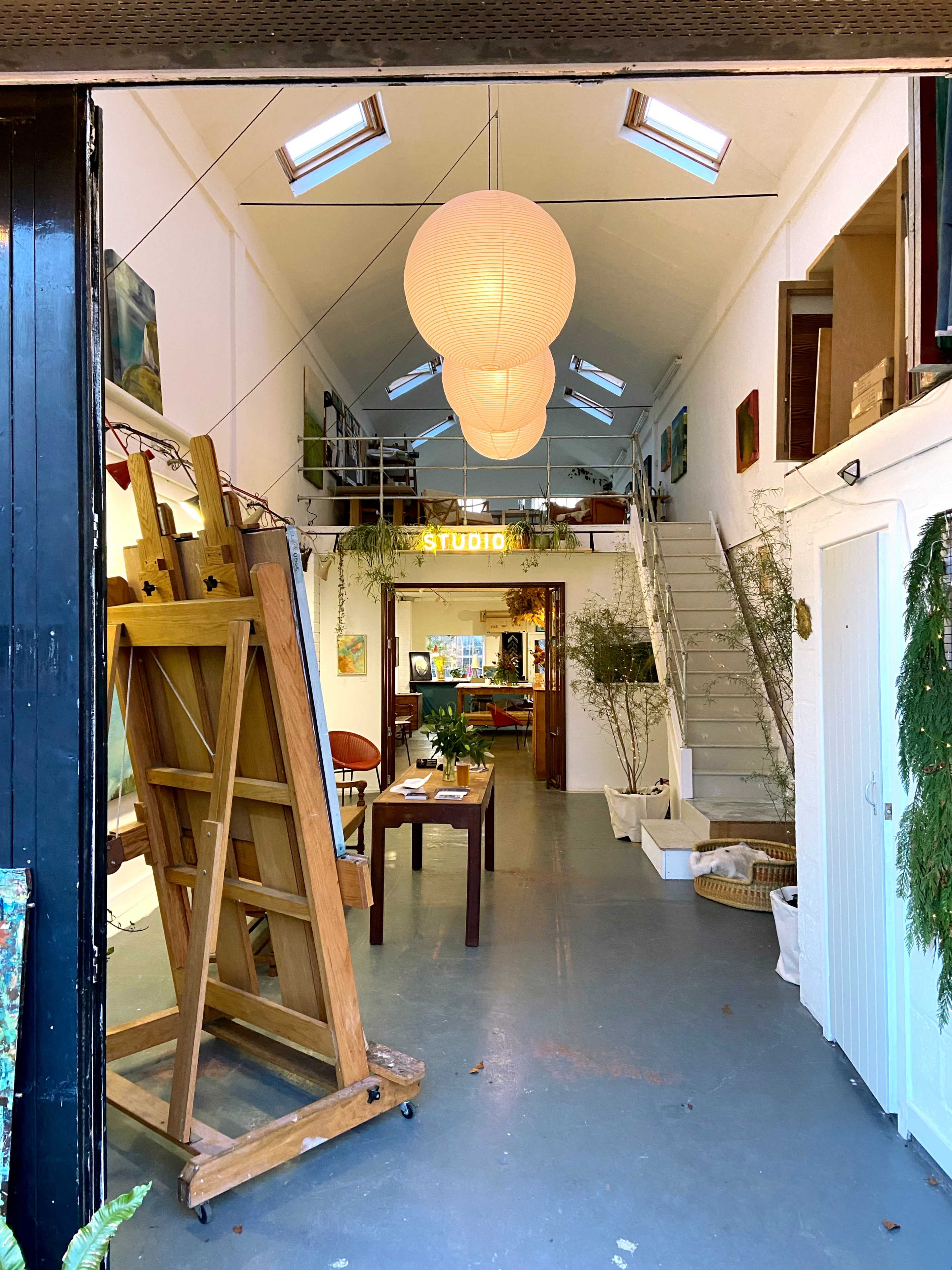The image shows the interior of an art studio with wooden easels, a table, and plants, illuminated by pendant lights and large windows.