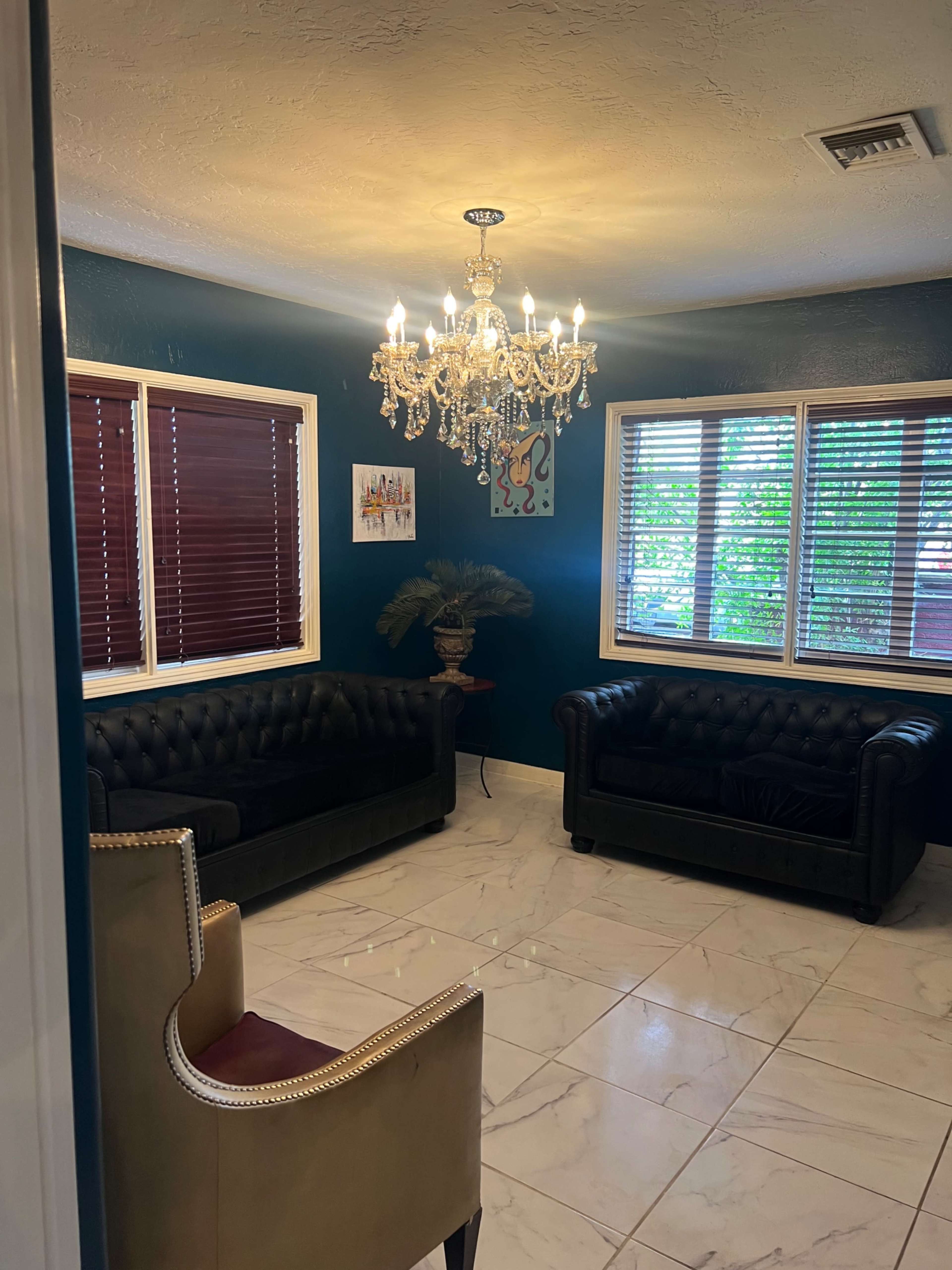 A well-lit sitting area features two black leather sofas, a chandelier, and windows with wooden blinds.