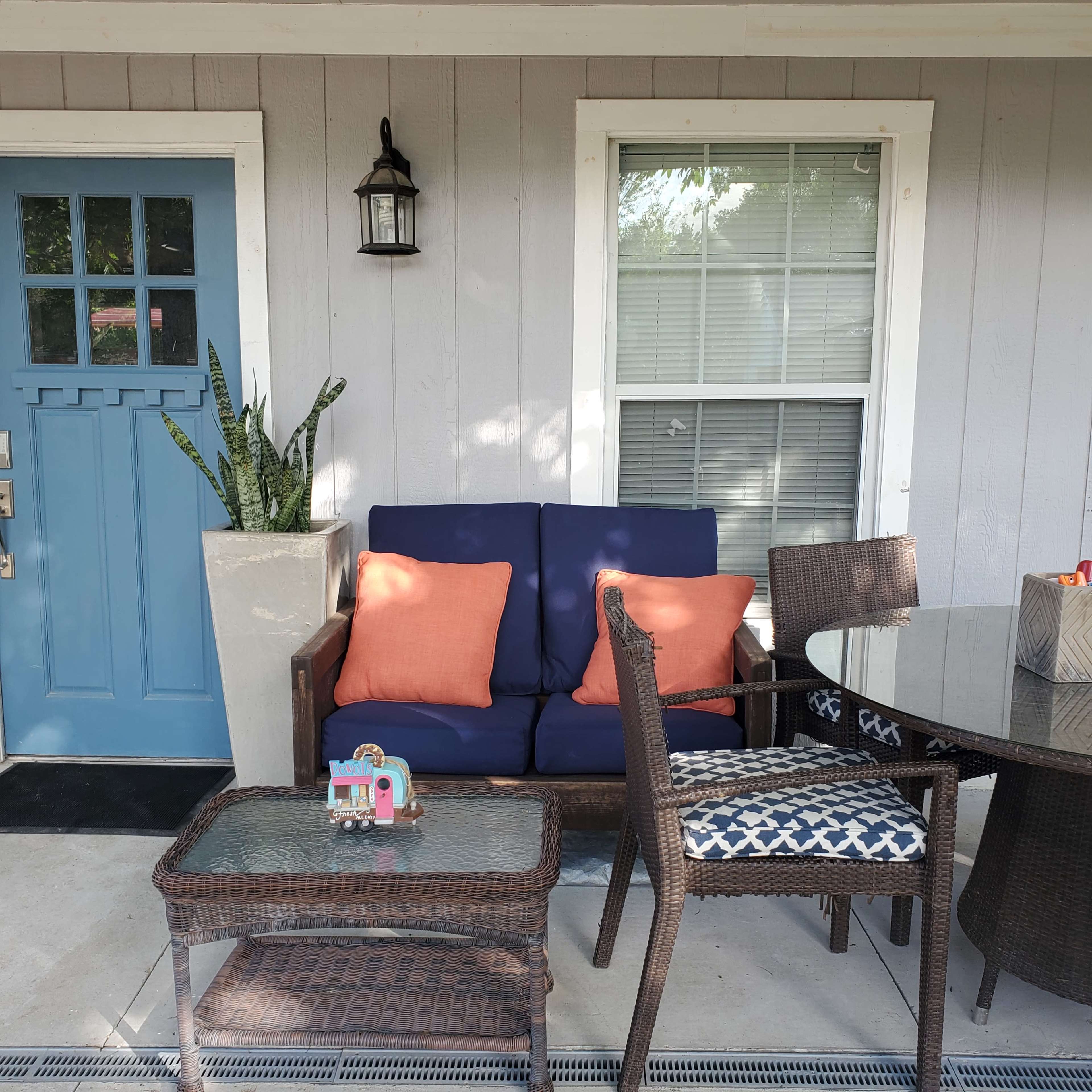A porch features a blue couch with orange pillows, a glass-topped table, and a wicker chair beside a potted plant.