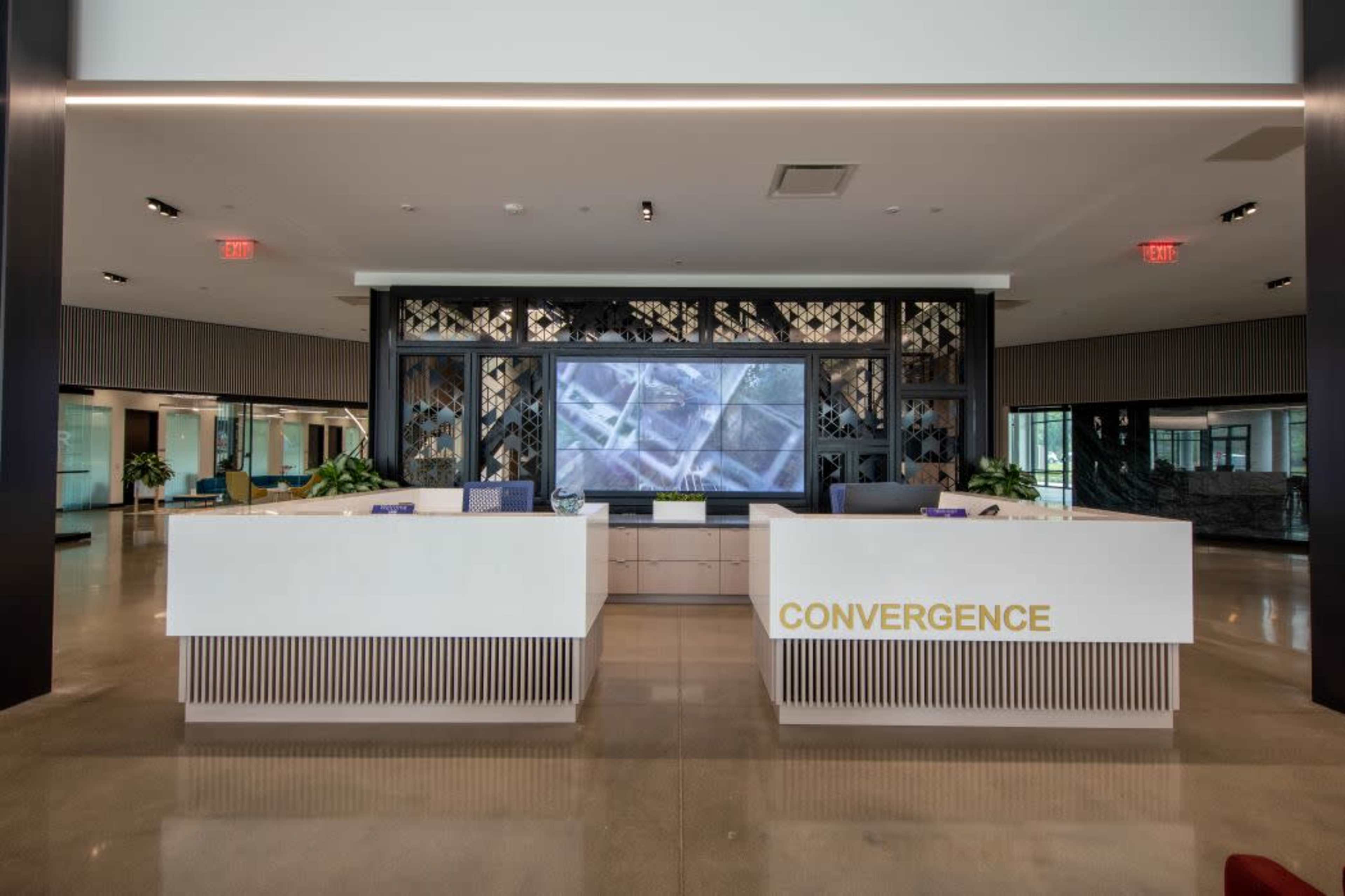 The image shows a modern reception area with two white desks labeled "CONVERGENCE" and a large screen displaying visuals in the background.