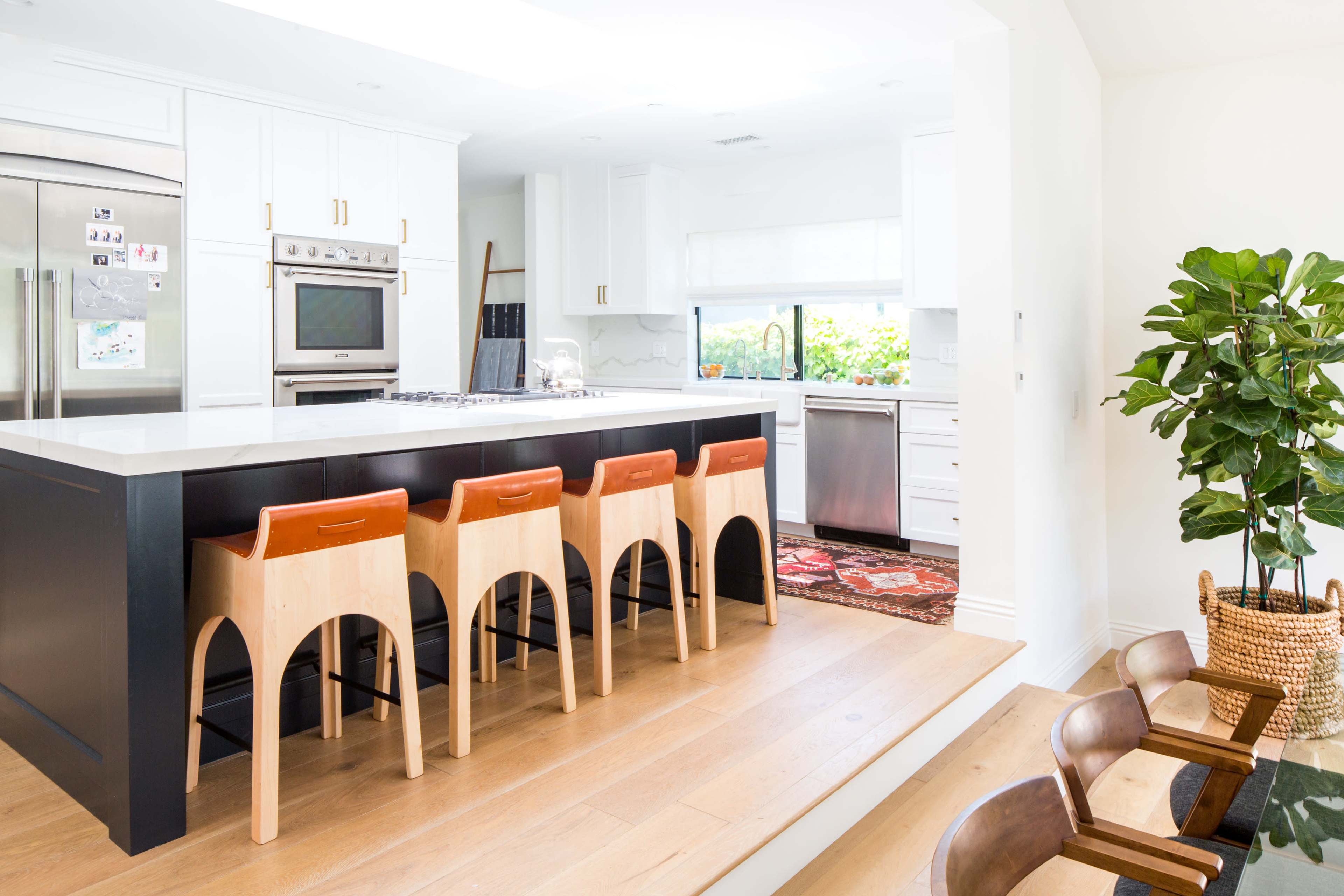 A modern kitchen features a large island with four wooden stools, stainless steel appliances, and abundant natural light.