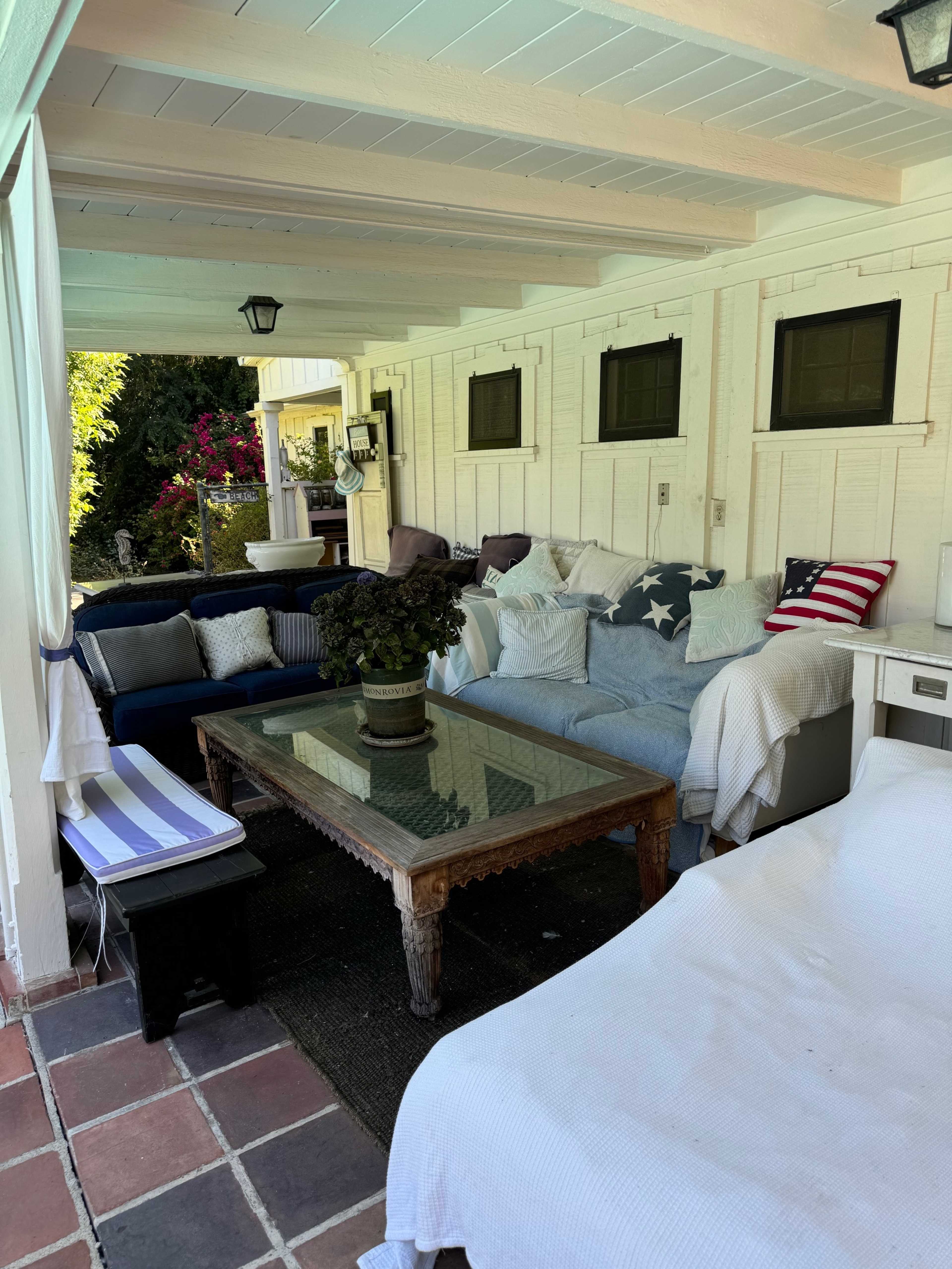 A covered patio area with several couches, a glass table in the center, and decorative plants.
