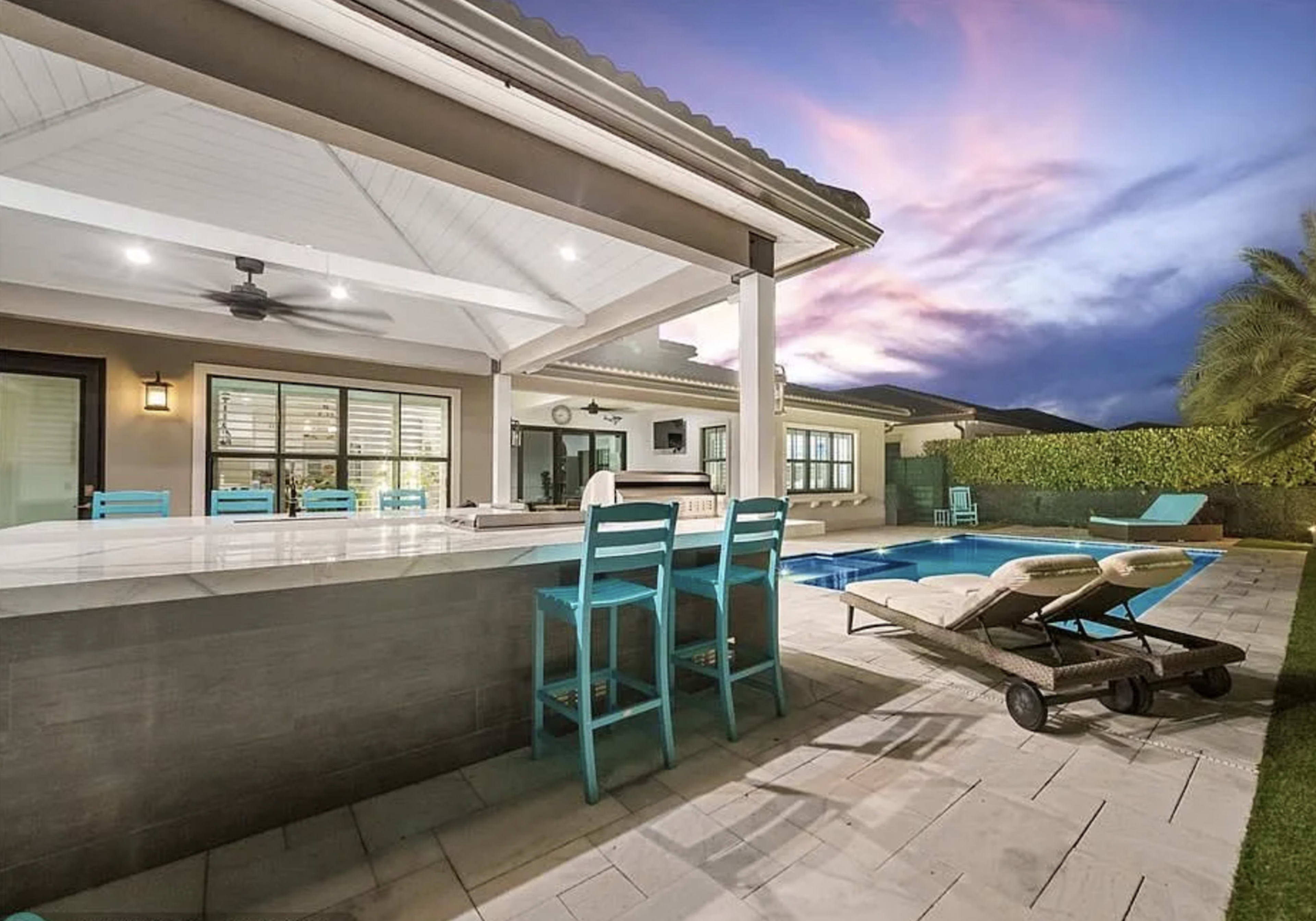 The image shows a modern outdoor patio with a swimming pool, lounge chairs, and a dining area featuring turquoise barstools under a covered roof.