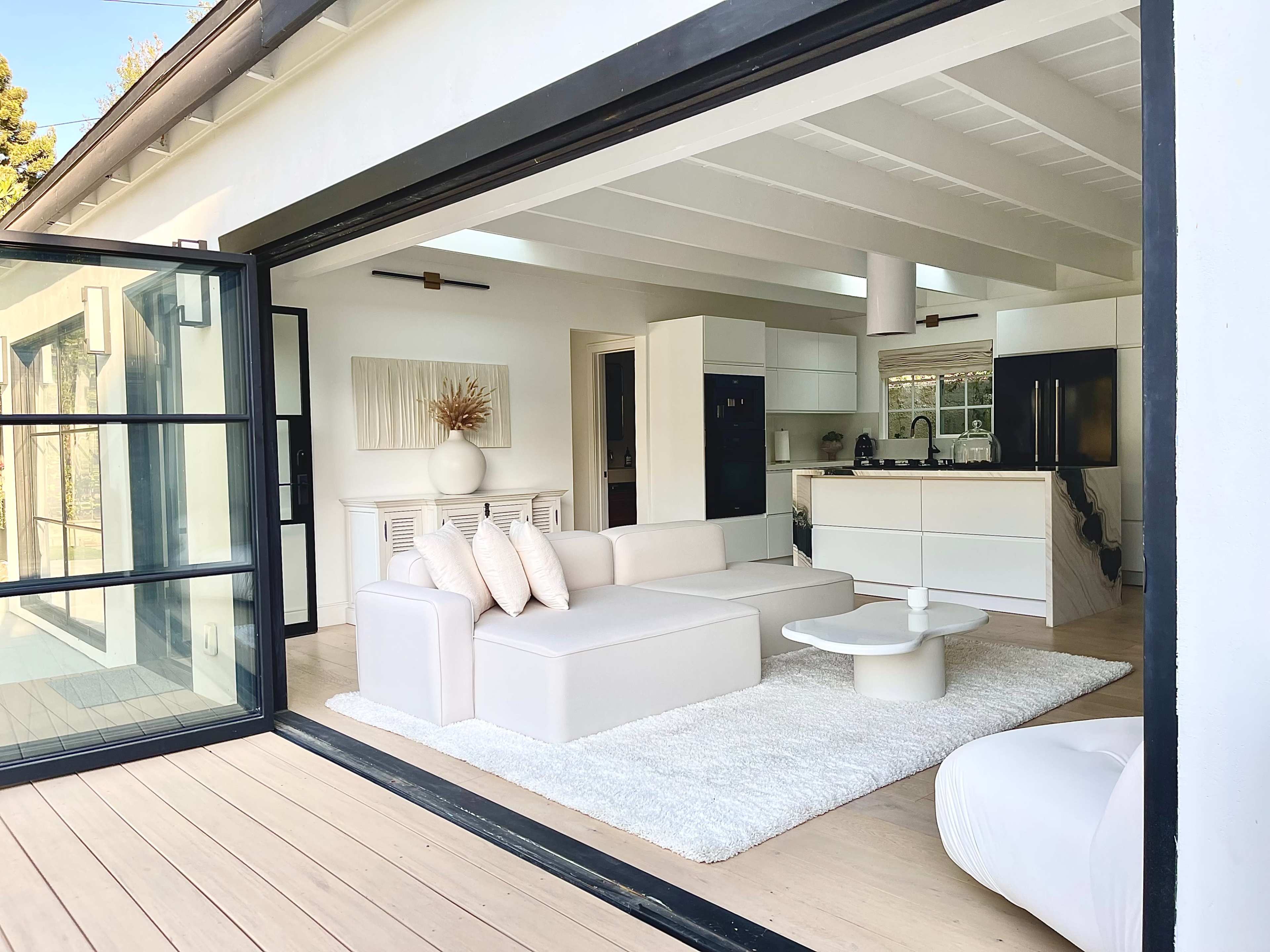 A bright, modern living space with a white sectional sofa, a coffee table, and a kitchen visible in the background through large open doors.