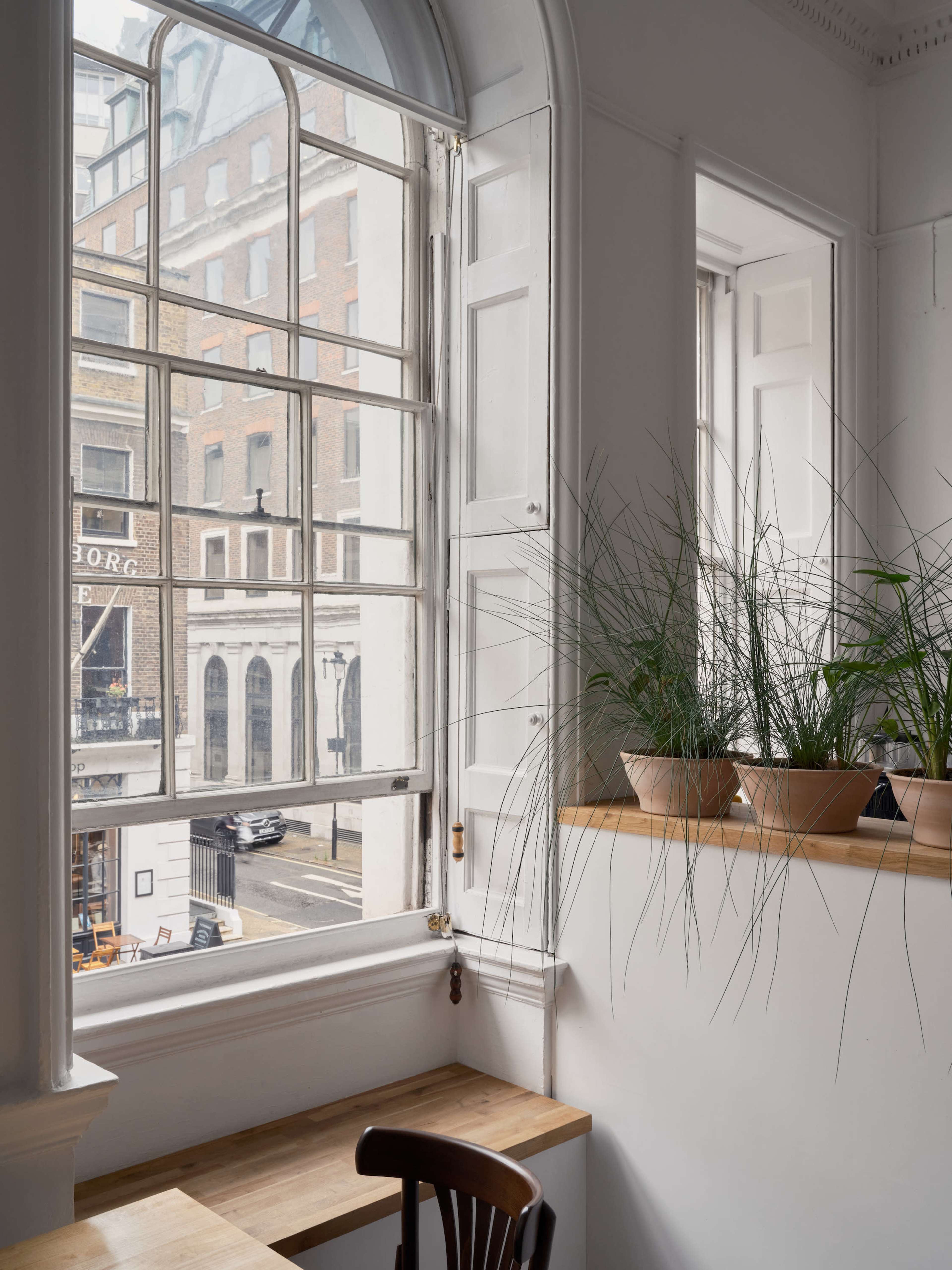 A well-lit interior room with large windows, wooden furniture, and potted plants on a ledge overlooking a street outside.