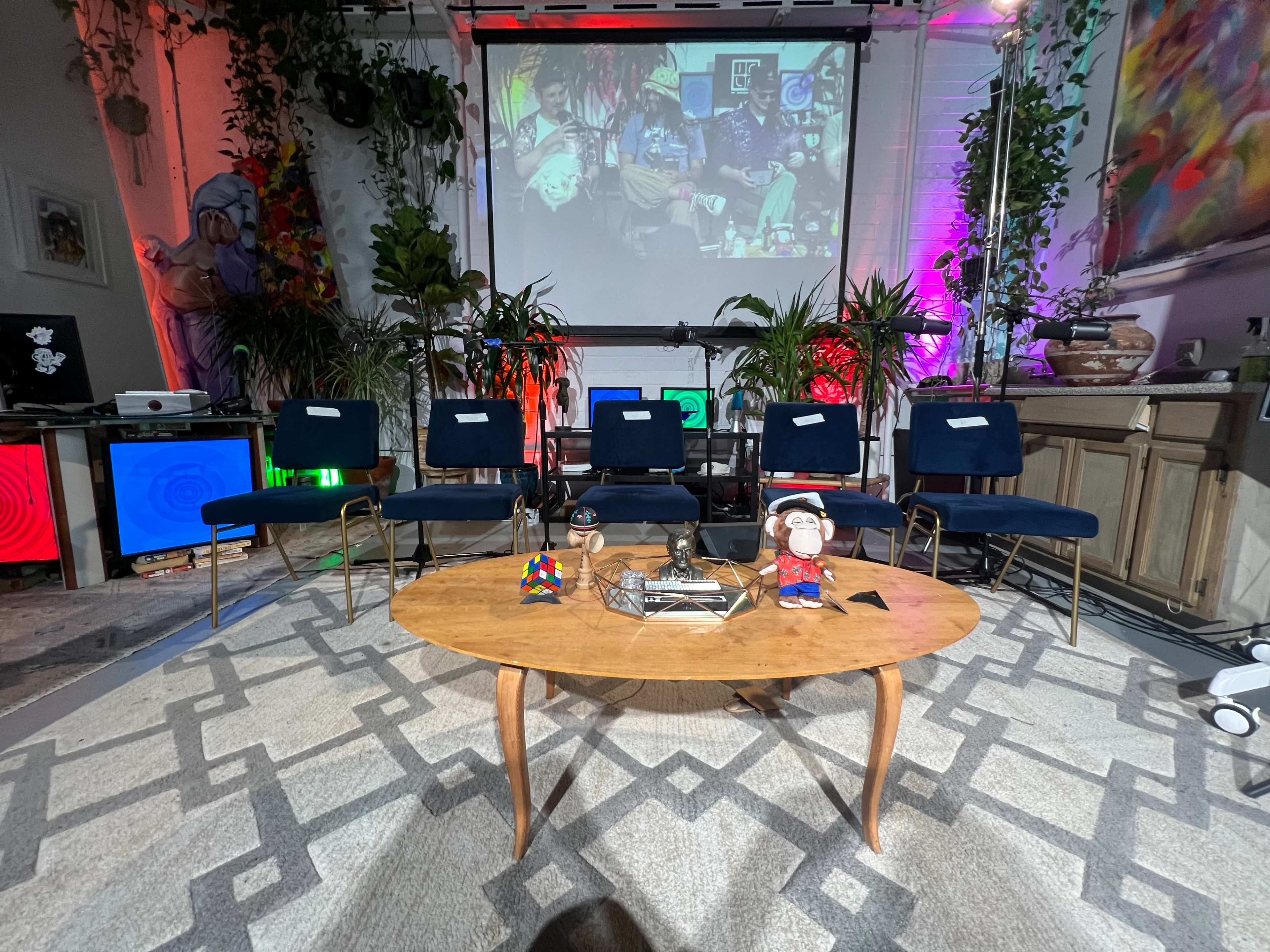 A set up for an event with a coffee table and several chairs arranged in front of a large screen displaying colorful artwork, surrounded by plants and decorative elements.