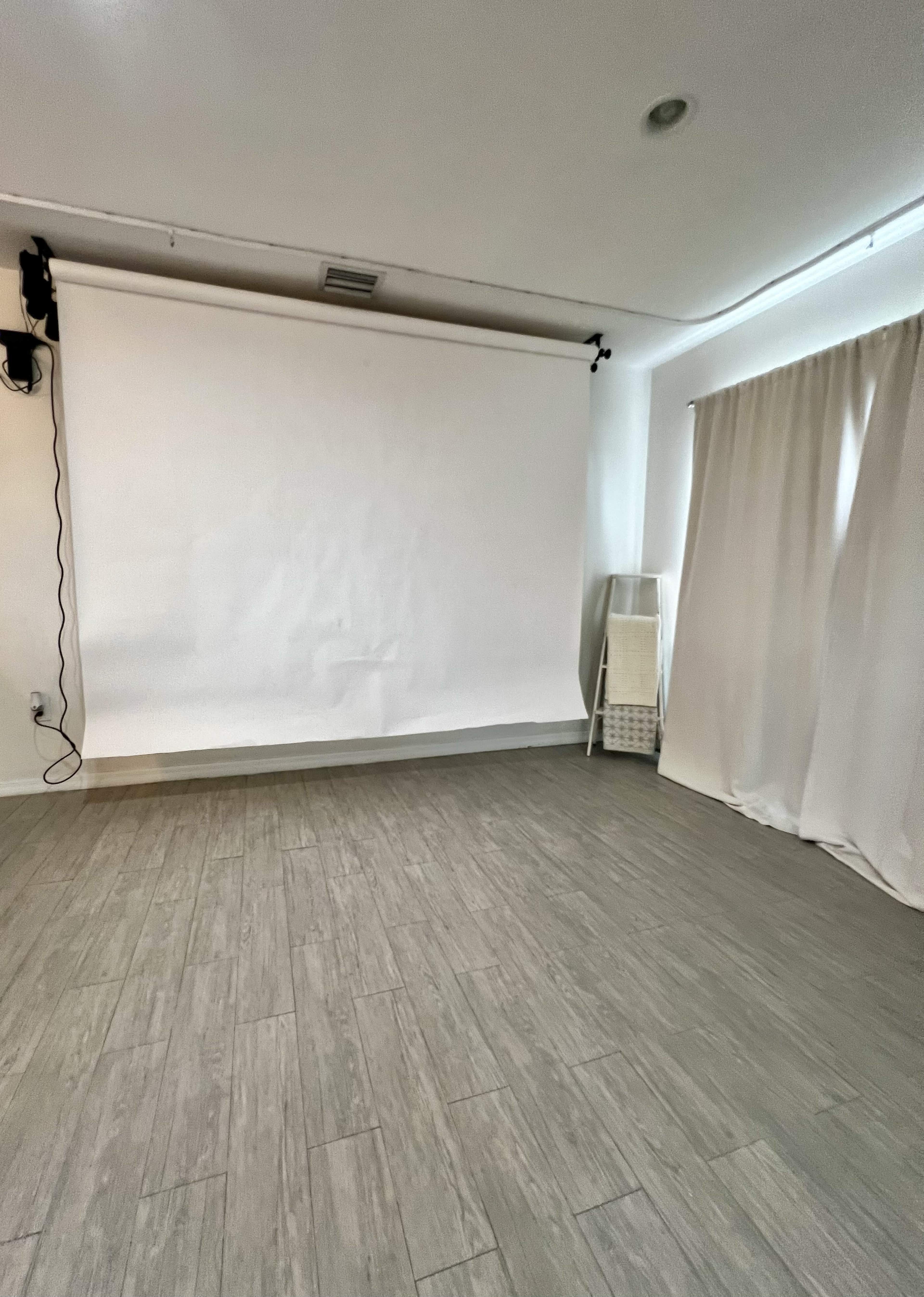 The image shows a room with a blank white projection screen mounted on the wall, a large window covered by a curtain, and a light-colored wooden floor.