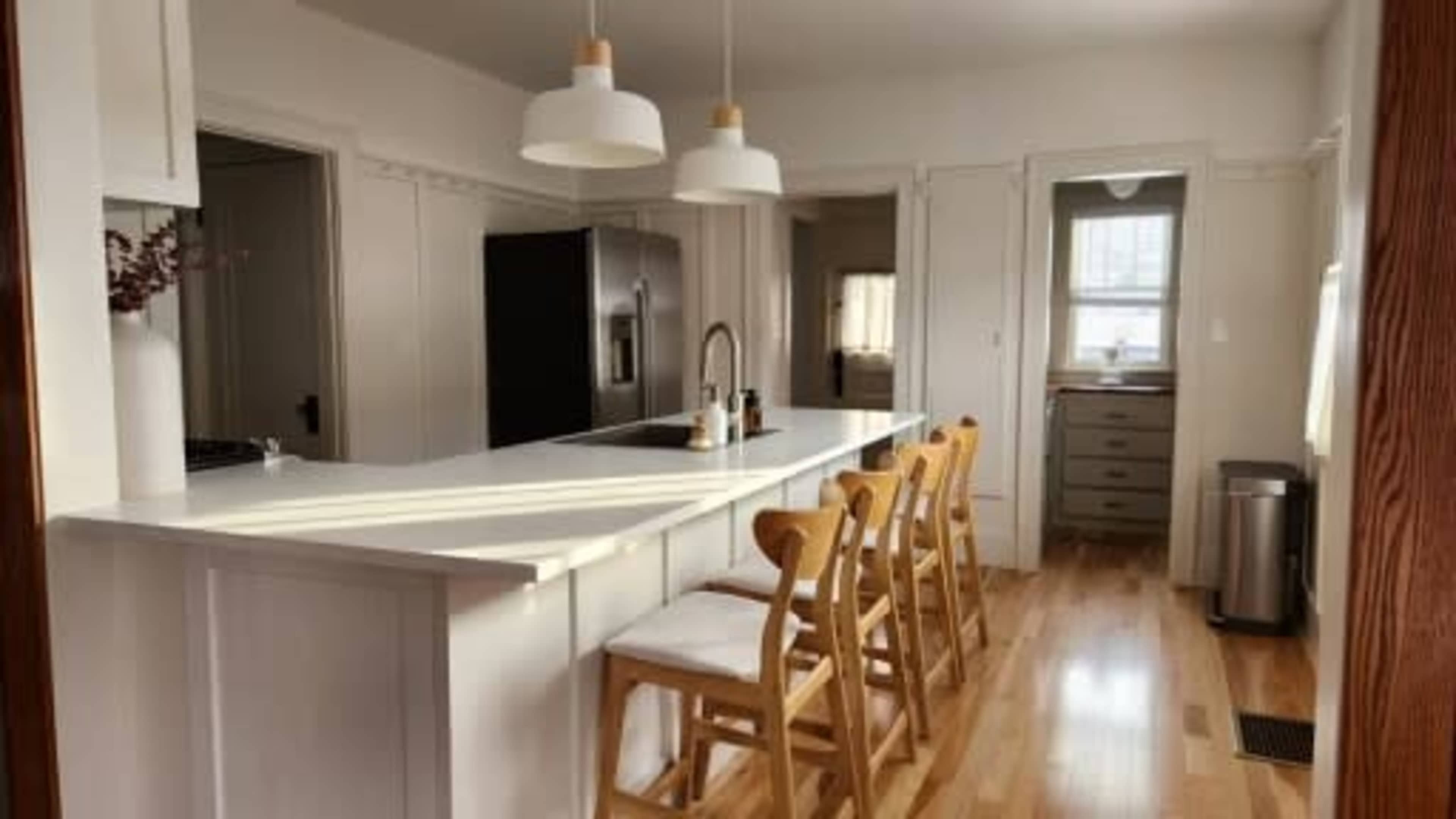 A modern kitchen features a long island with six wooden chairs, a black refrigerator, and large pendant lights hanging above.