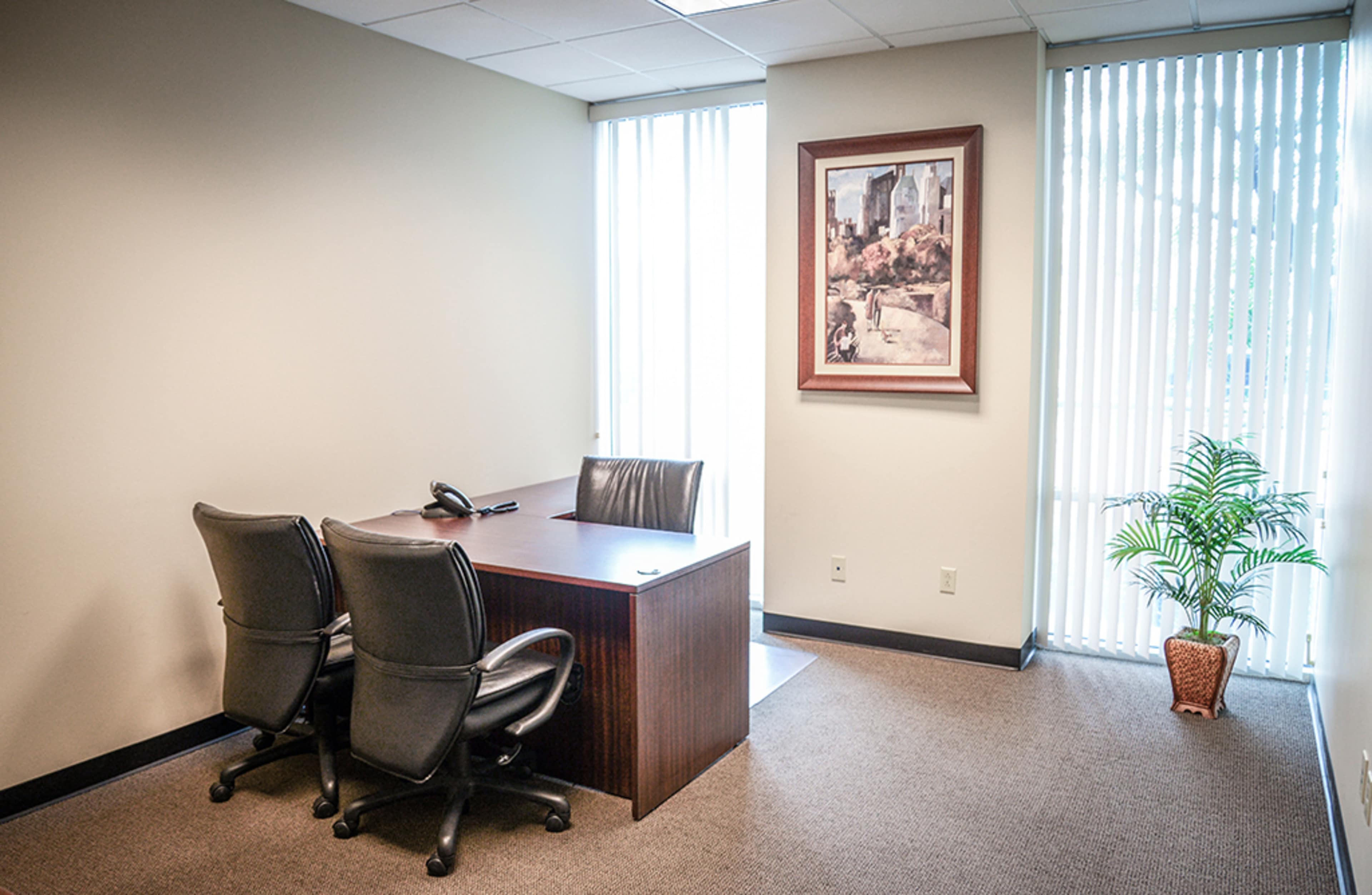 A simple office space with a wooden desk, two black chairs, a telephone, and a framed painting on the wall, along with a plant in a pot.