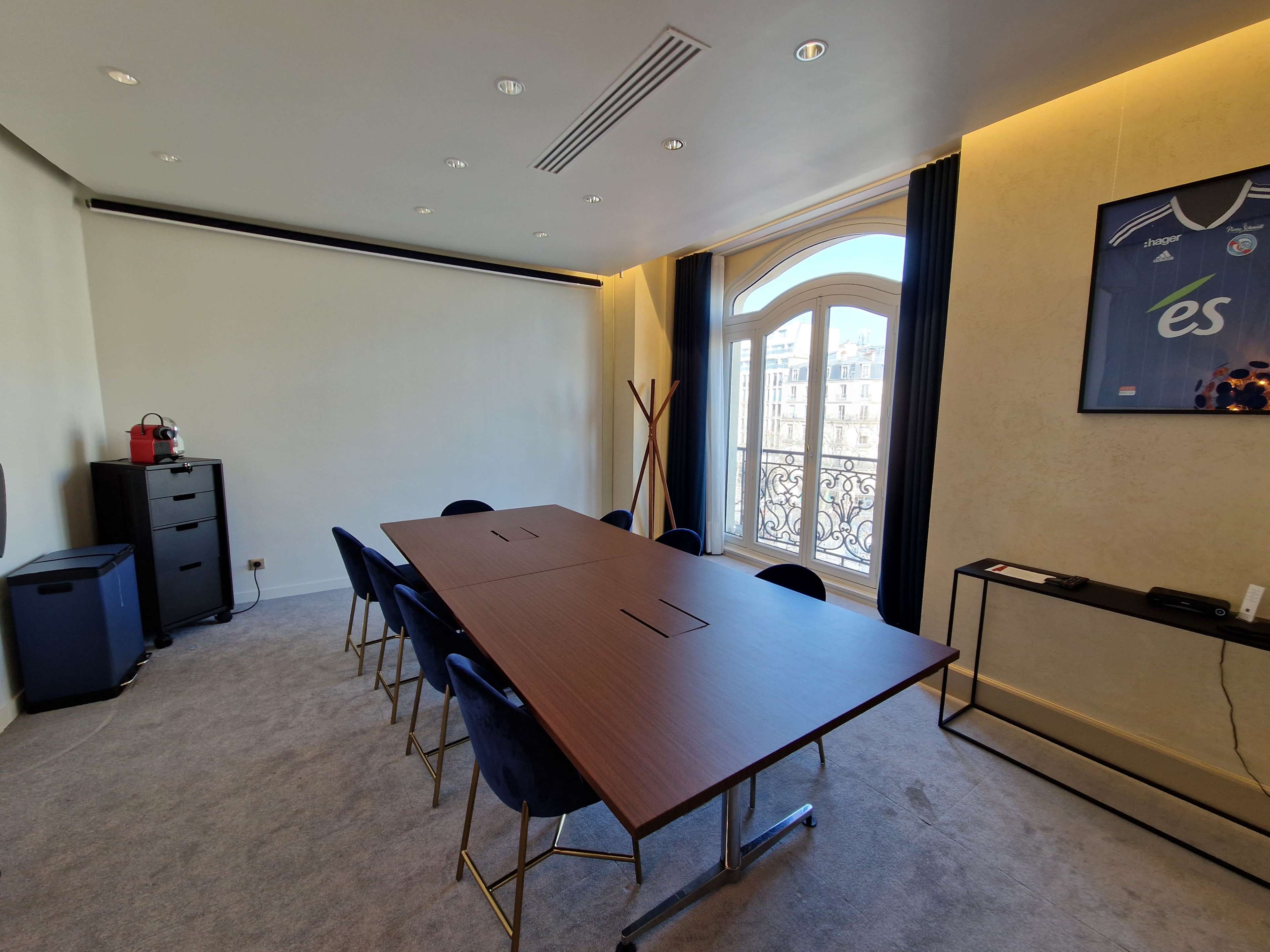 A modern meeting room features a long wooden table surrounded by blue chairs, with a large window offering a view of the city outside.