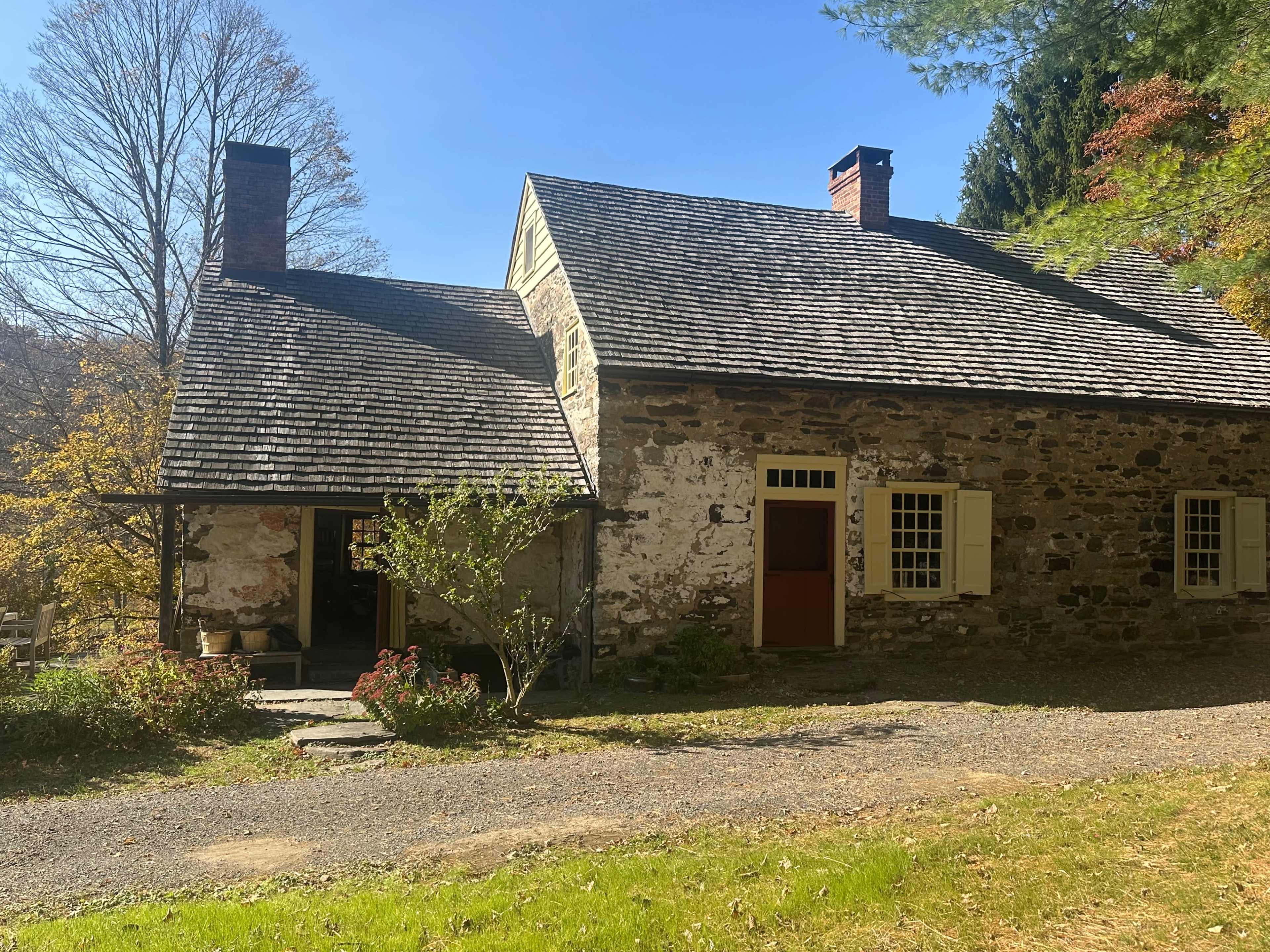 The image shows a historic stone house with a slanted roof, set beside a gravel path and surrounded by trees.