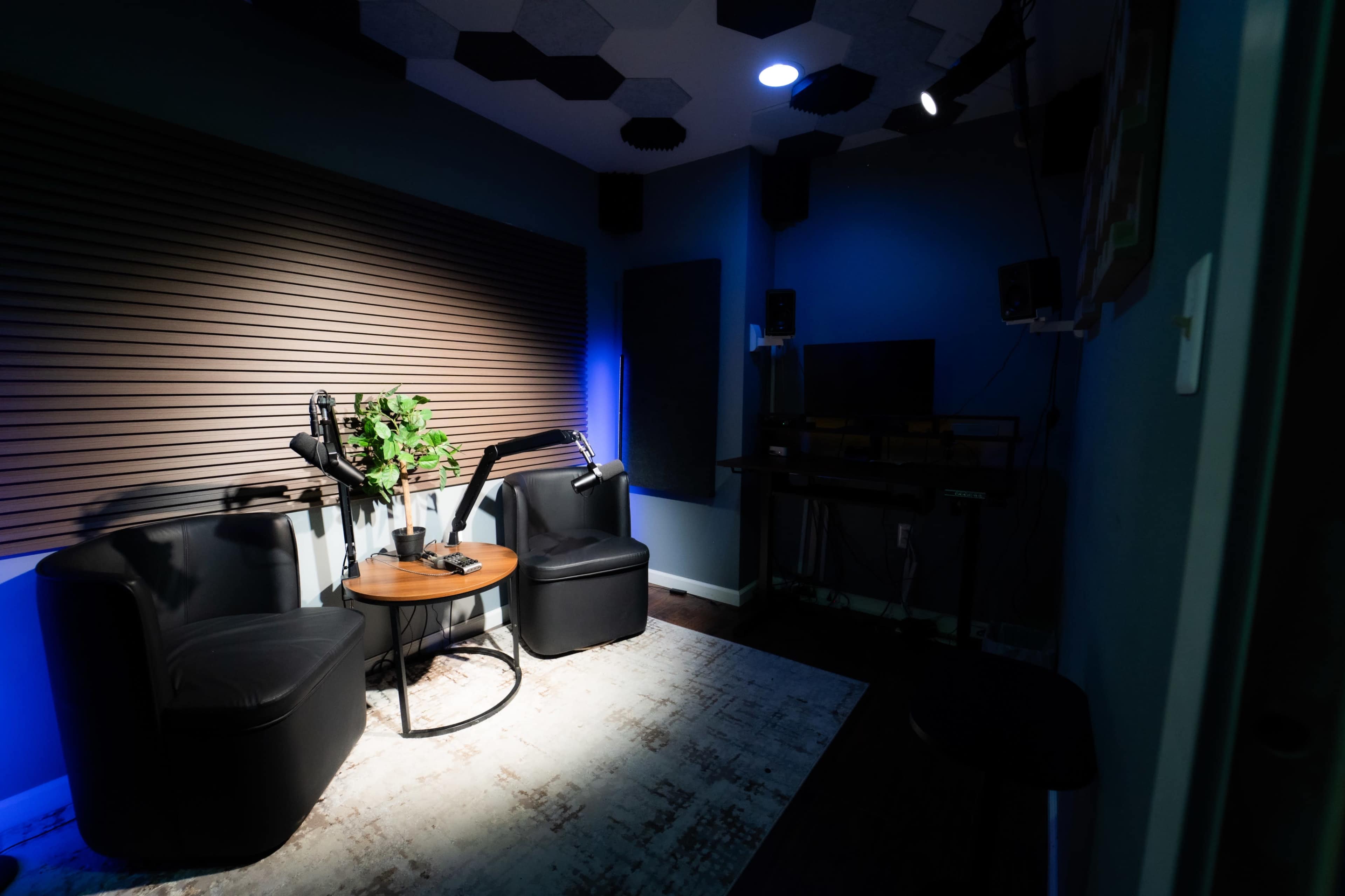 The image shows a small recording studio with two black chairs, a round table, and soundproof walls, illuminated by blue lighting.