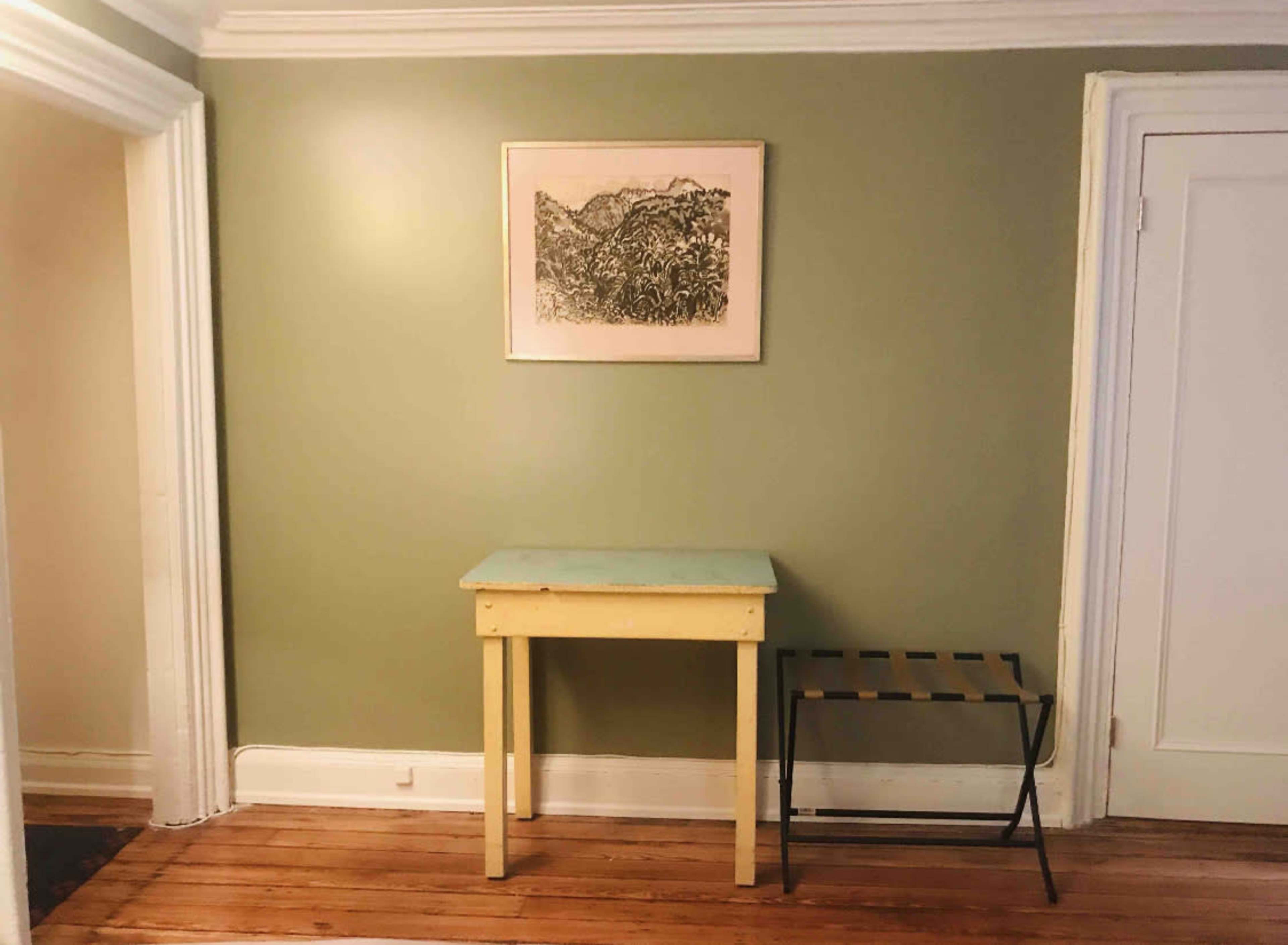A small yellow table with a green top and a black metal stand is positioned against a olive-green wall with a framed artwork above it.